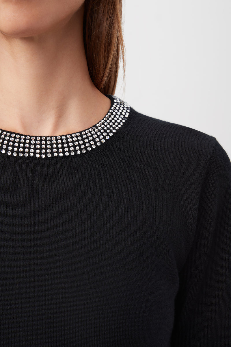 Close-up of a person wearing the Laelia Merino Wool Sweater With Rhinestone Trim, featuring a round neckline decorated with two rows of small, shiny rhinestones; only the lower face, neck, and upper chest are visible.