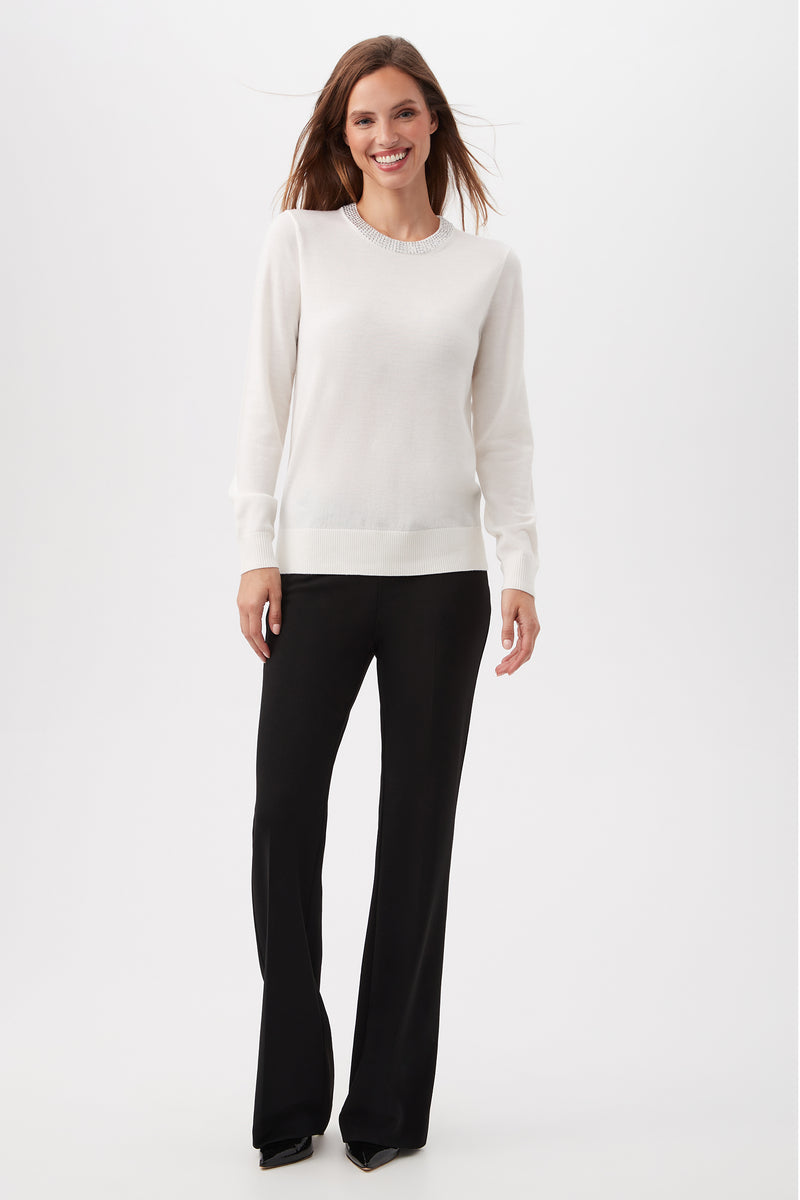 A woman stands smiling against a plain white background, wearing the Laelia Merino Wool Sweater With Rhinestone Trim paired with black dress pants and black shoes.