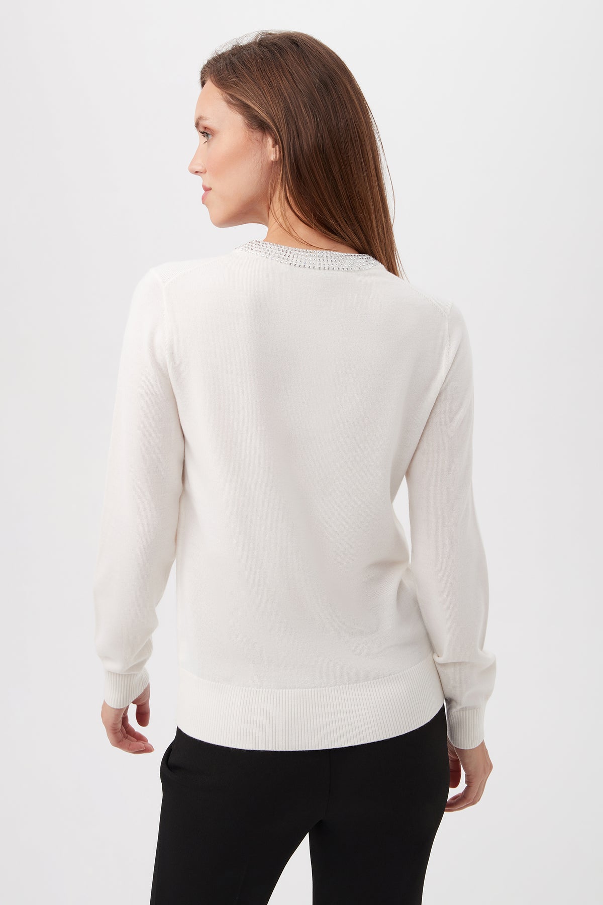 A woman with long brown hair stands turned to the side, showcasing the back of the Laelia Merino Wool Sweater With Rhinestone Trim and black pants against a plain light background.