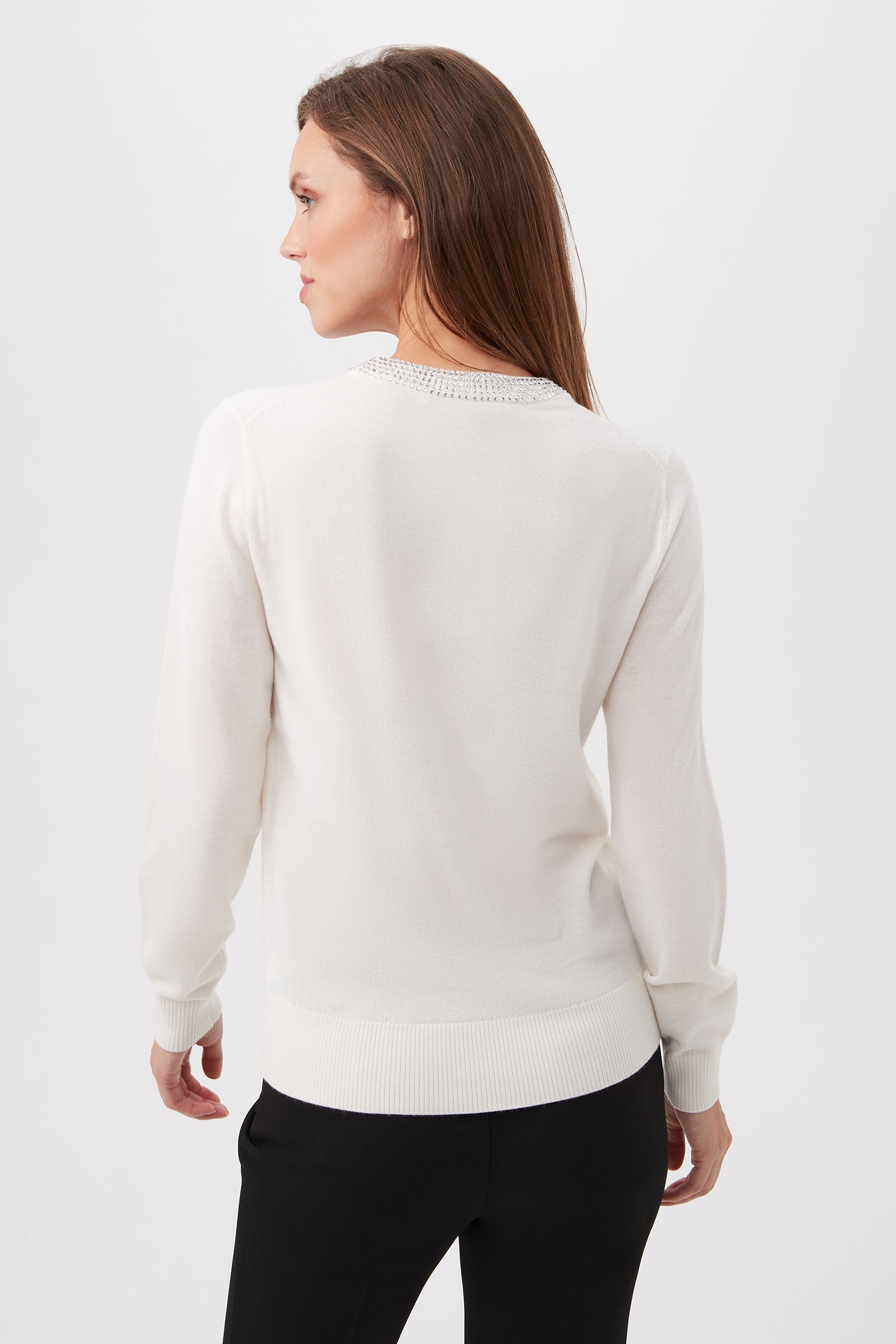 A woman with long brown hair stands turned to the side, showcasing the back of the Laelia Merino Wool Sweater With Rhinestone Trim and black pants against a plain light background.