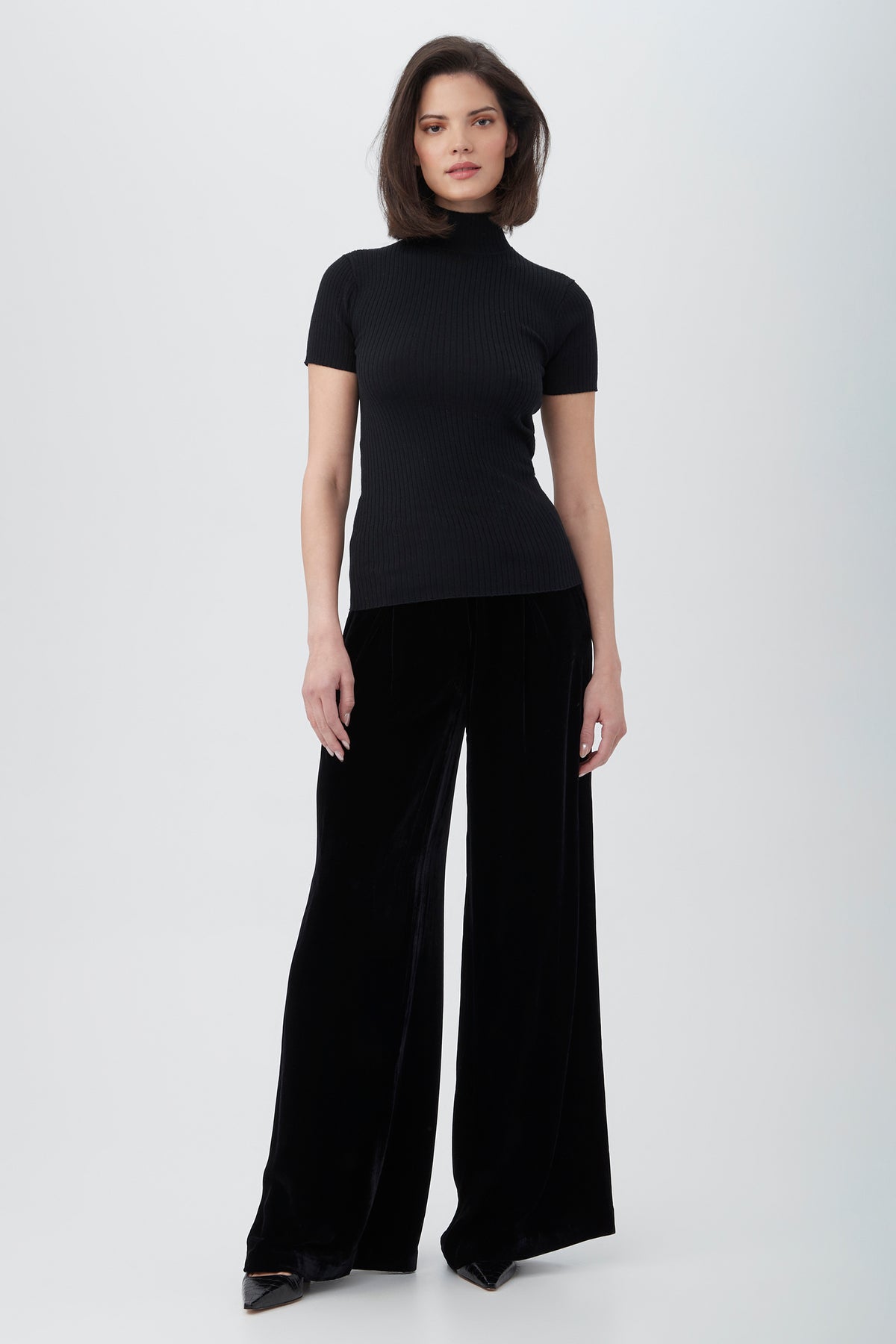 A woman with a bob hairstyle and neutral expression stands against a light background, wearing black pointed shoes, a fitted black tee, and Saige Velvet Wide Leg Pants.