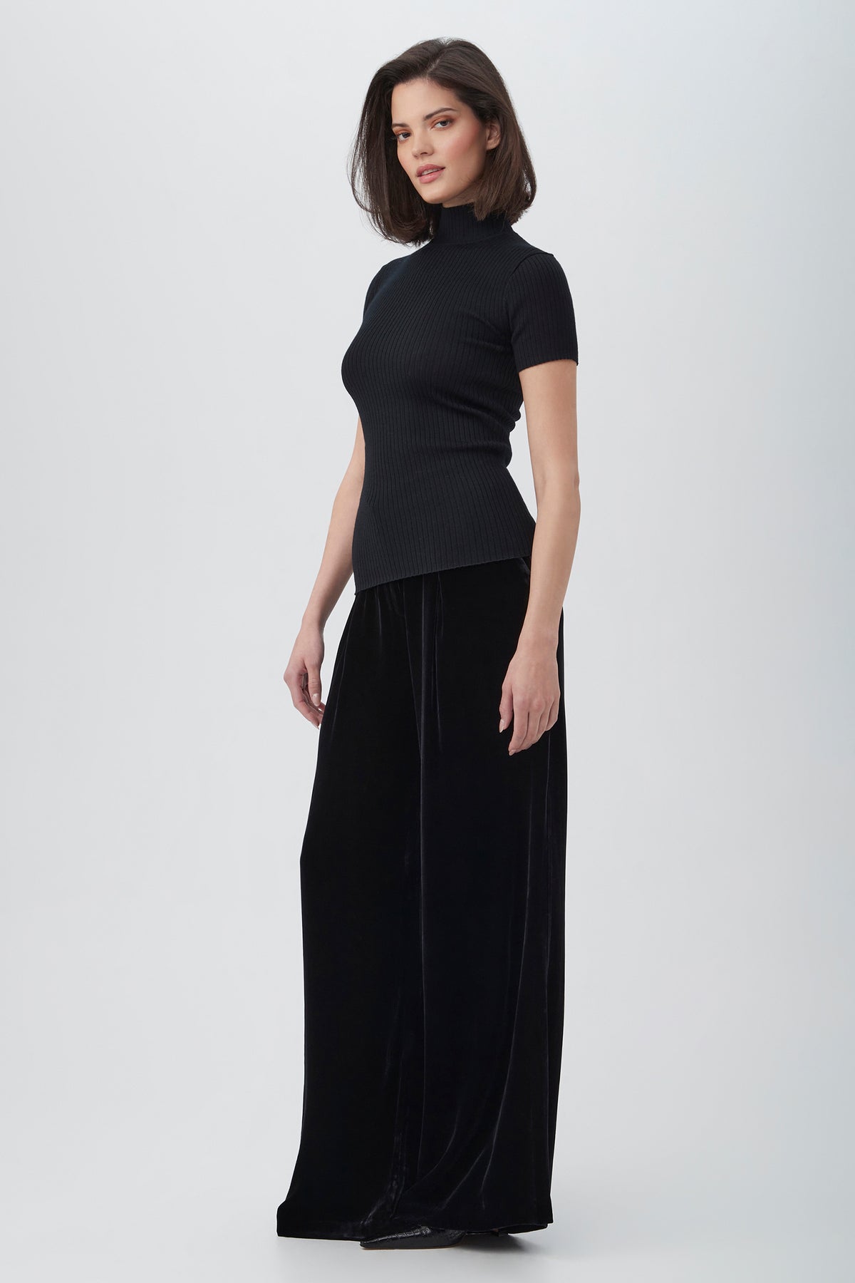 A woman with straight, shoulder-length dark hair and a neutral expression stands against a plain light background, wearing the Saige Velvet Wide Leg Pants in black with a matching short-sleeve turtleneck top.