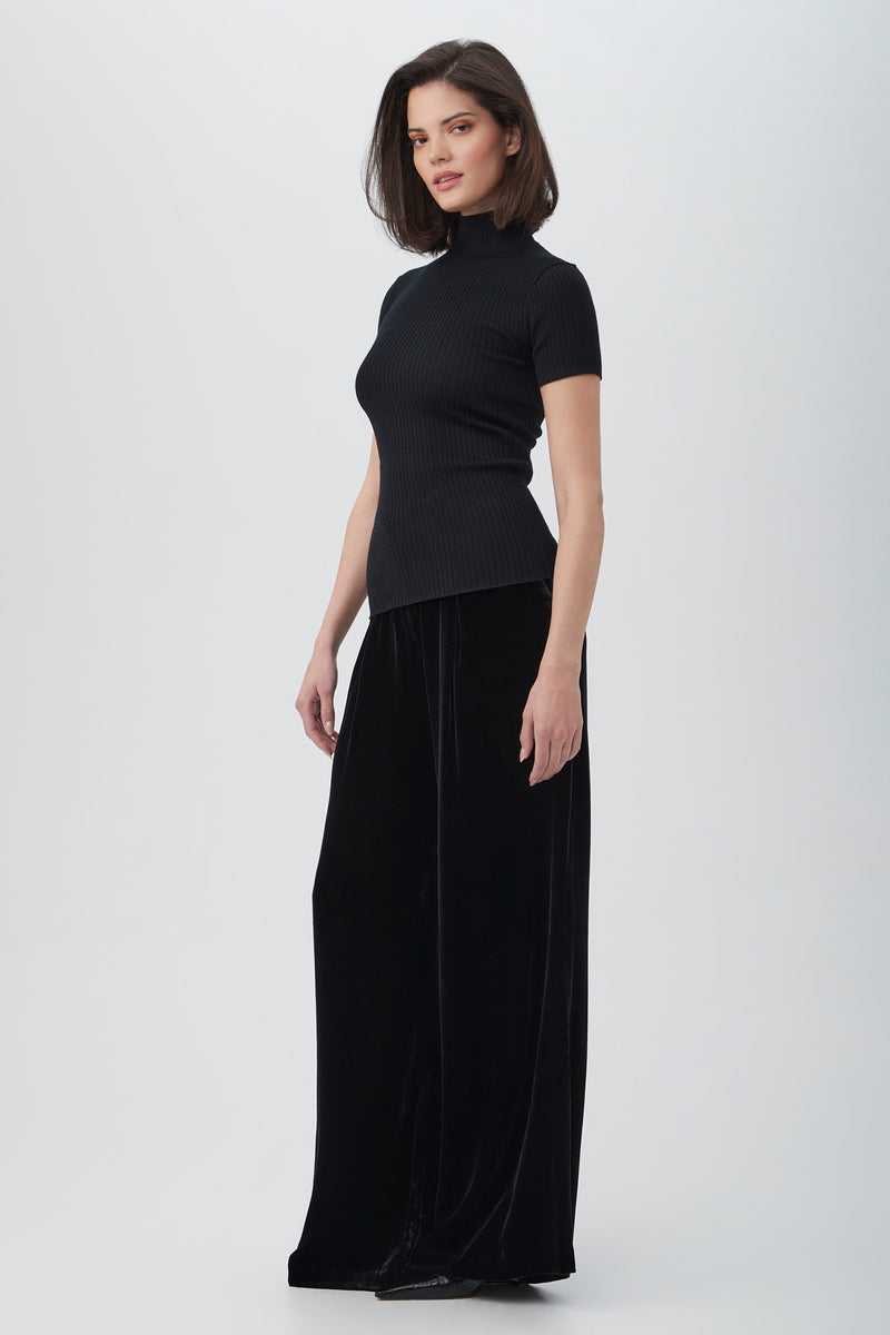 A woman with straight, shoulder-length dark hair and a neutral expression stands against a plain light background, wearing the Saige Velvet Wide Leg Pants in black with a matching short-sleeve turtleneck top.