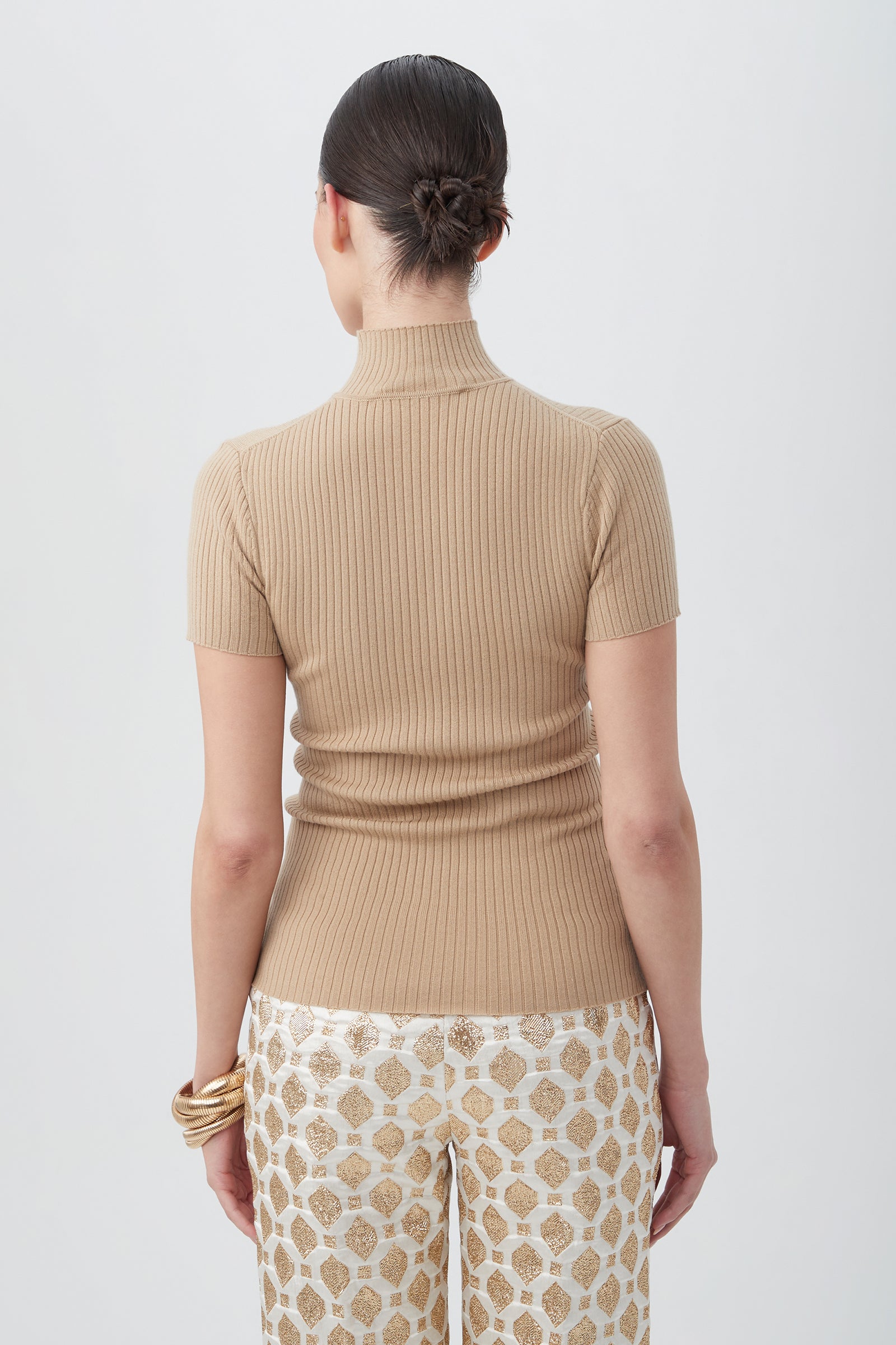 A woman with dark hair in a low bun, wearing the Armeni Merino Wool Short Sleeve Mockneck Top and patterned cream and gold pants, stands facing away against a plain light background.