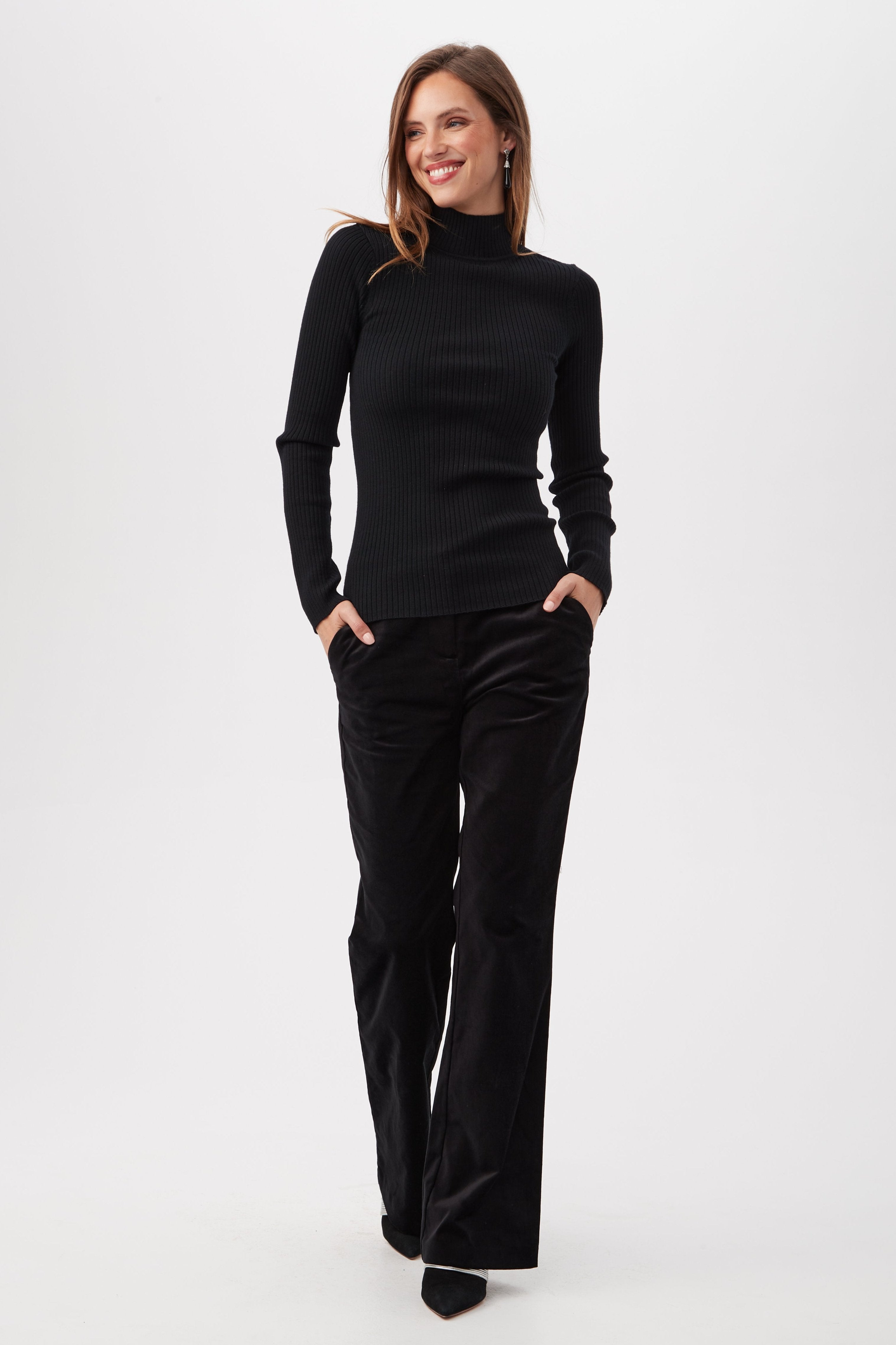 A woman smiles with her hands in her pockets, wearing the Sade Merino Wool Mockneck Sweater With Open Back and loose black pants against a plain white background.