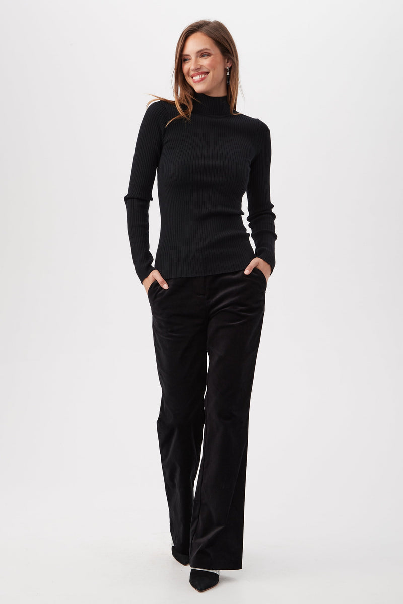 A woman smiles with her hands in her pockets, wearing the Sade Merino Wool Mockneck Sweater With Open Back and loose black pants against a plain white background.