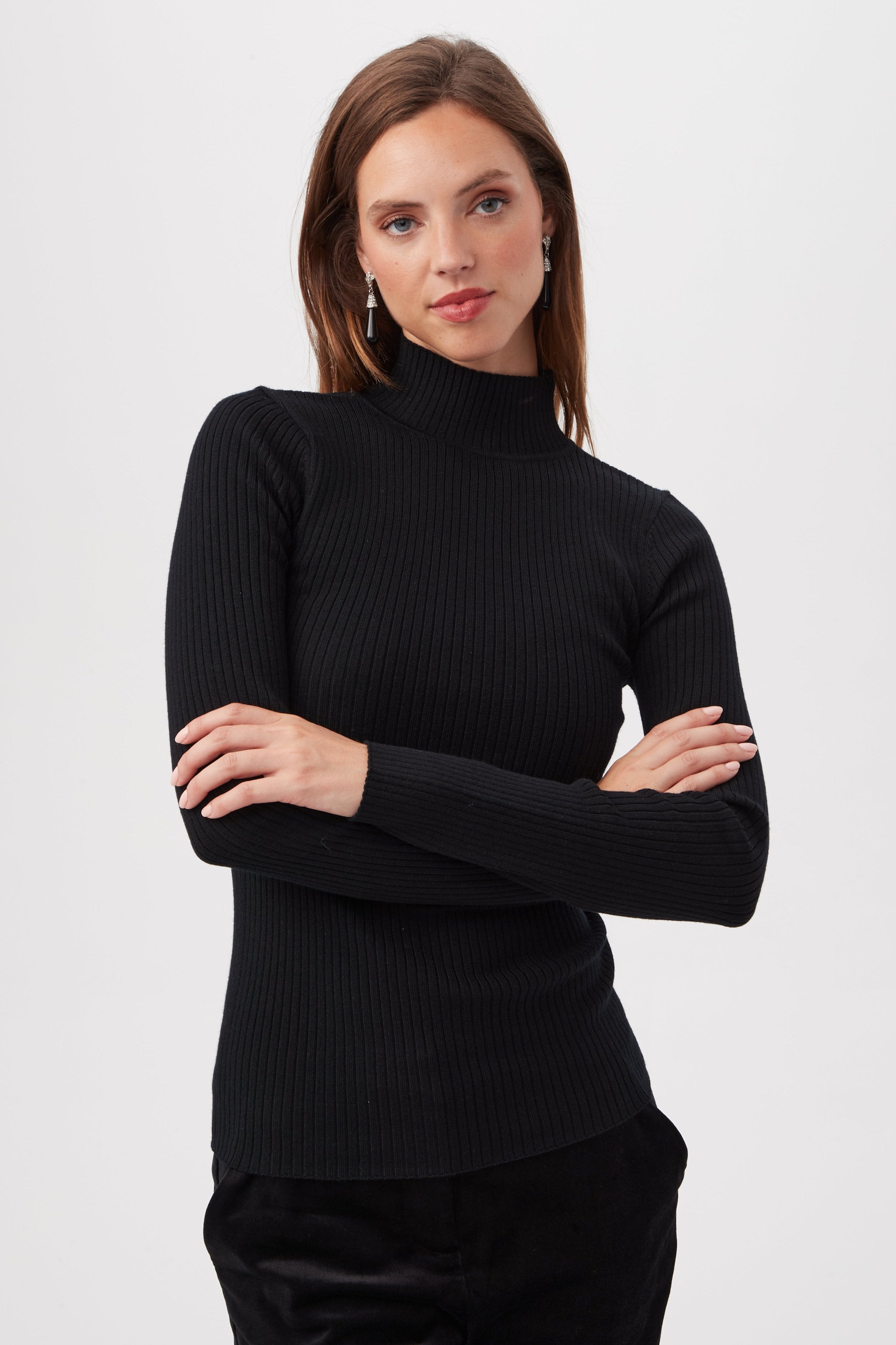 A woman with long brown hair wears the Sade Merino Wool Mockneck Sweater With Open Back and silver earrings, standing confidently with arms crossed against a plain white background.