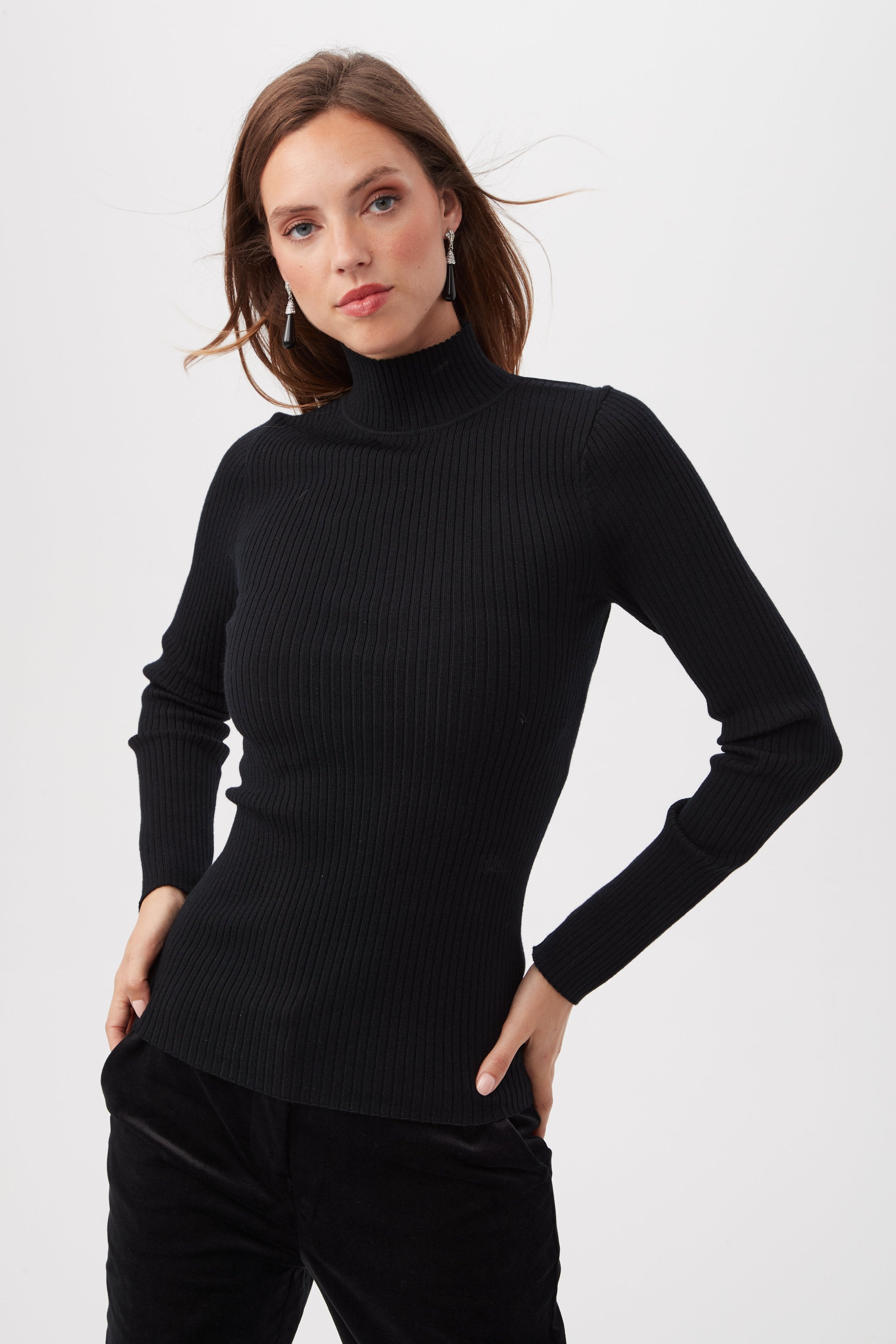 A woman with straight brown hair wears the Sade Merino Wool Mockneck Sweater With Open Back in black and black pants, standing with her hands on her hips against a plain white background.