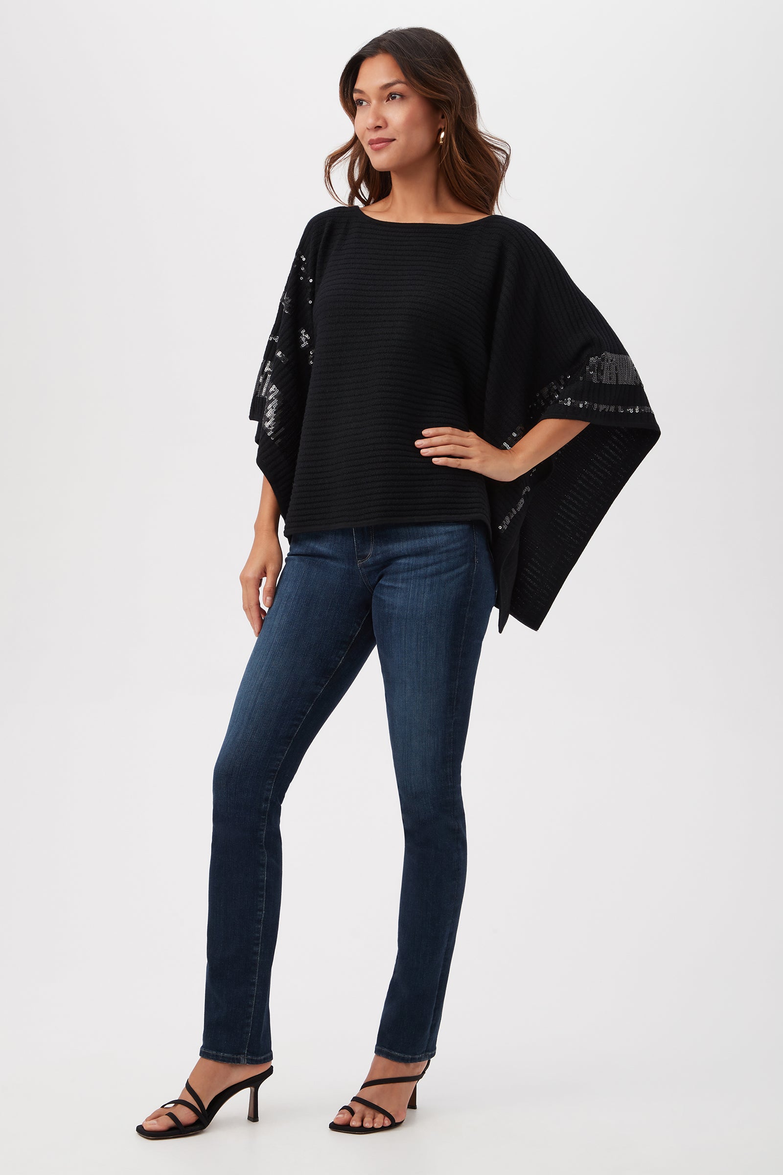 A woman poses in the Adela Merino Wool Wrap Sweater With Black Sequin Stripes, paired with dark blue skinny jeans and black heeled sandals. She has long brown hair and stands against a plain white background.