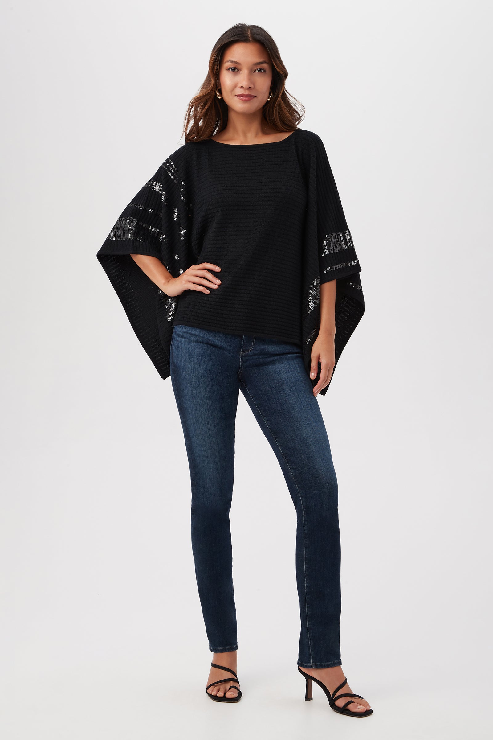 A woman with long hair and a confident stance, hand on her hip, models the Adela Merino Wool Wrap Sweater With Black Sequin Stripes against a white background, paired with fitted blue jeans and black heeled sandals.