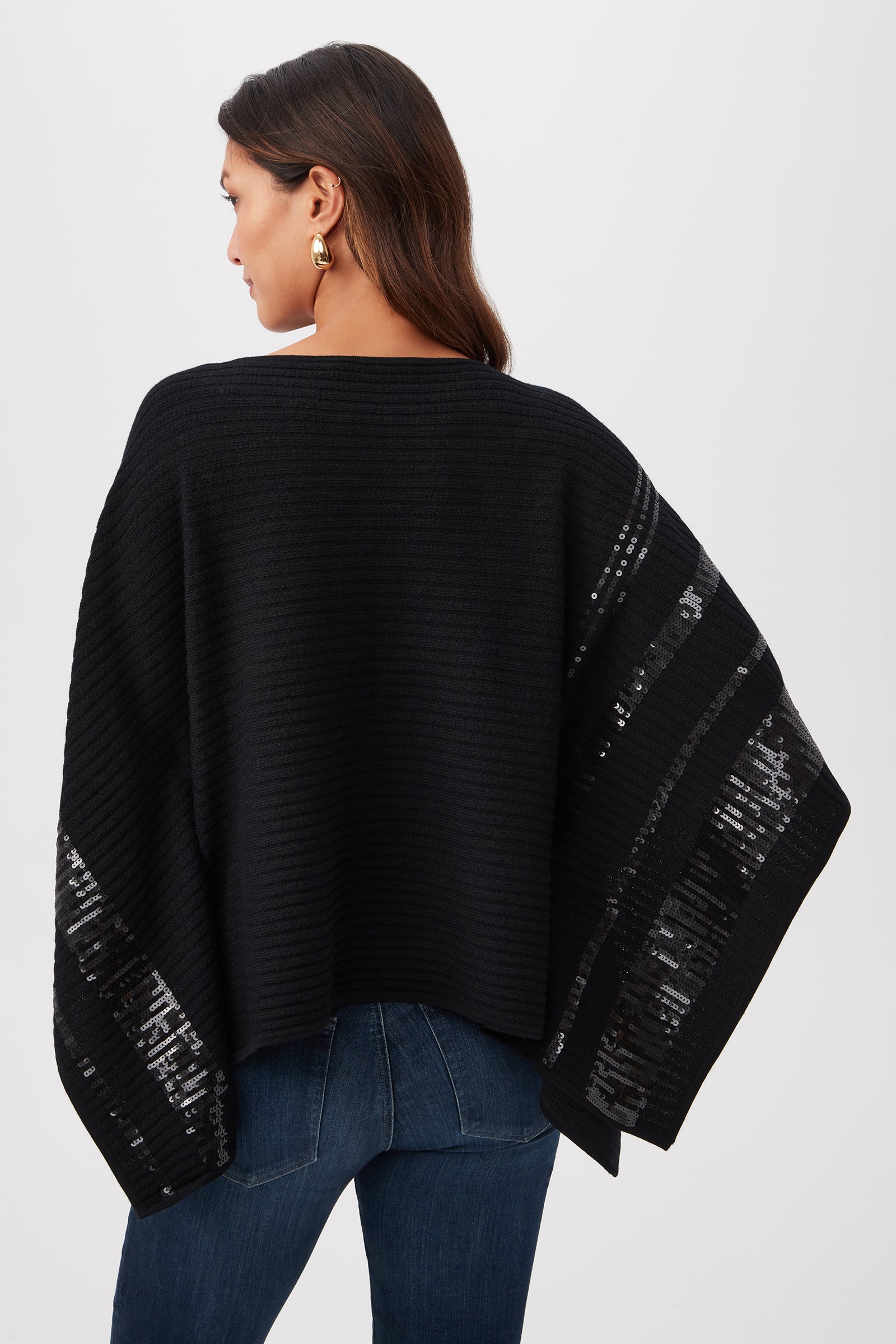 A woman with long brown hair and gold earrings faces away, wearing the Adela Merino Wool Wrap Sweater with black sequin stripes and blue jeans, set against a plain light background.