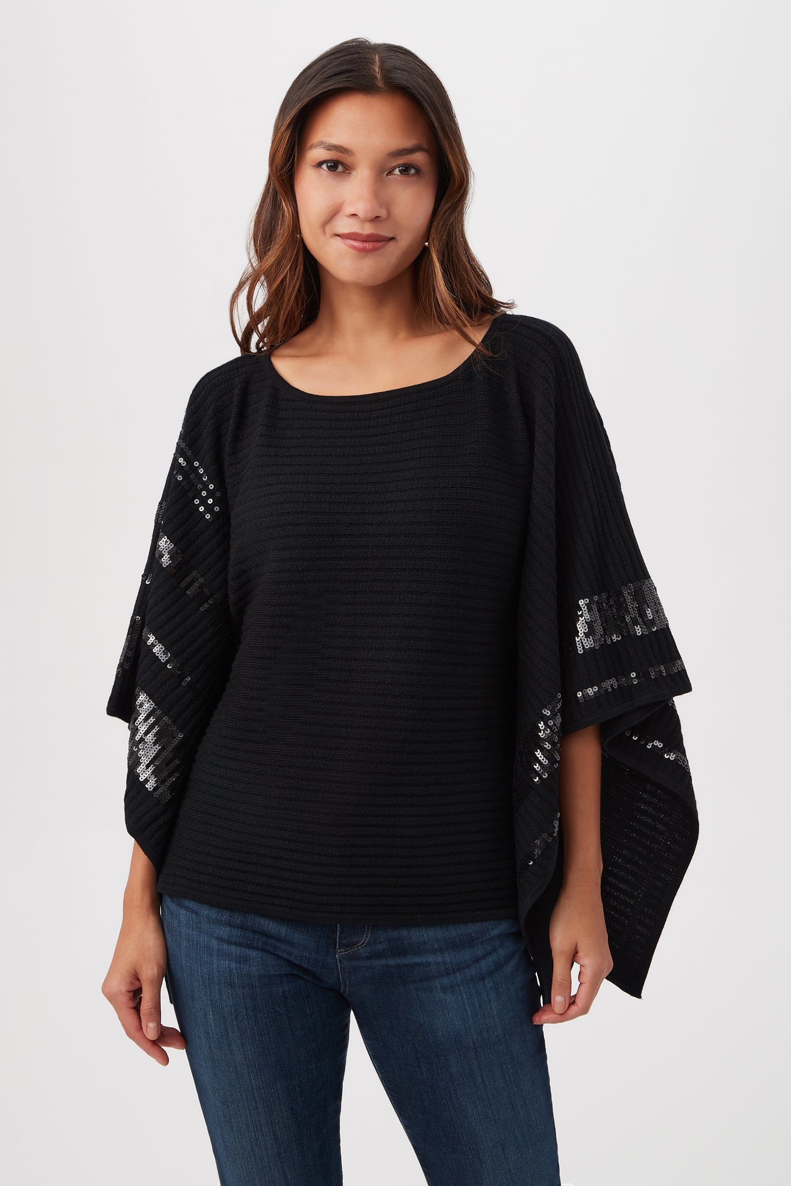 A woman with long brown hair models the Adela Merino Wool Wrap Sweater in black, featuring black sequin stripes on the sleeves, paired with blue jeans against a plain white background.