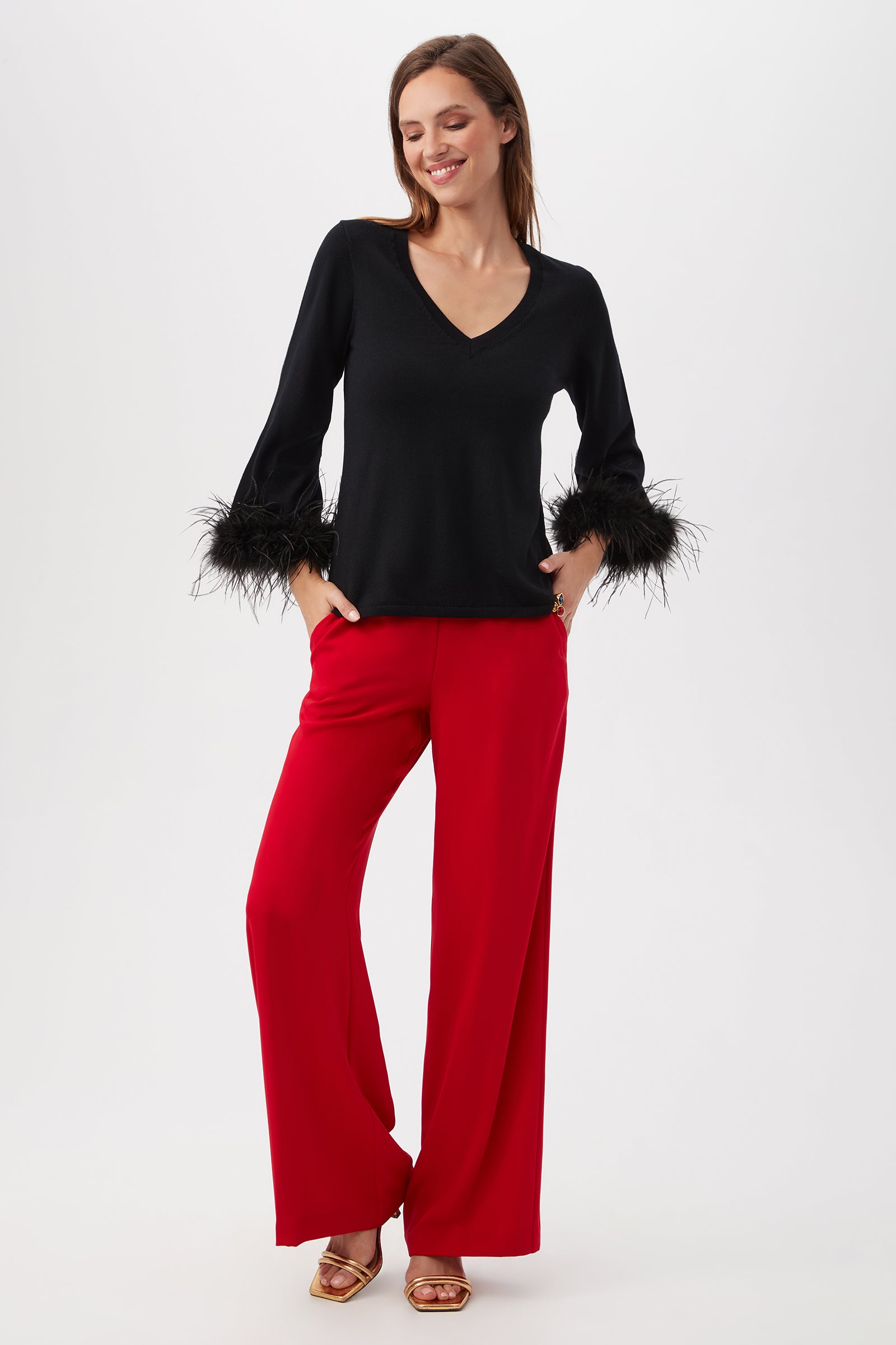 Woman standing and smiling, wearing the Sassy Merino Wool Feather Trimmed Sweater in black with feathered cuffs, paired with wide-leg red pants and gold open-toe heels against a plain white background.