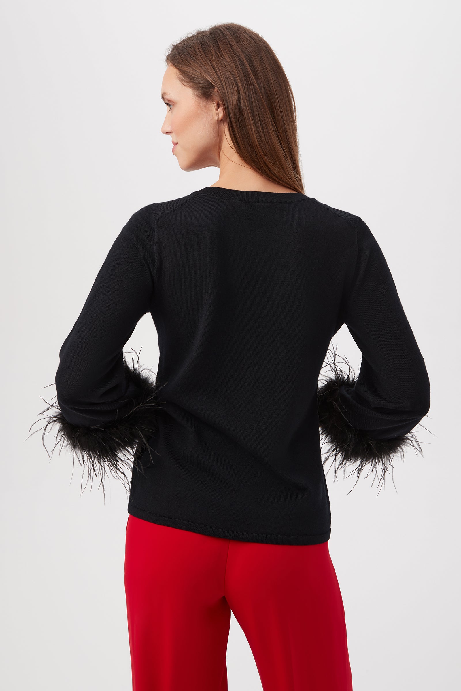 A woman with straight brown hair faces away, wearing the Sassy Merino Wool Feather Trimmed Sweater in black with feather cuffs and red pants, set against a light background.