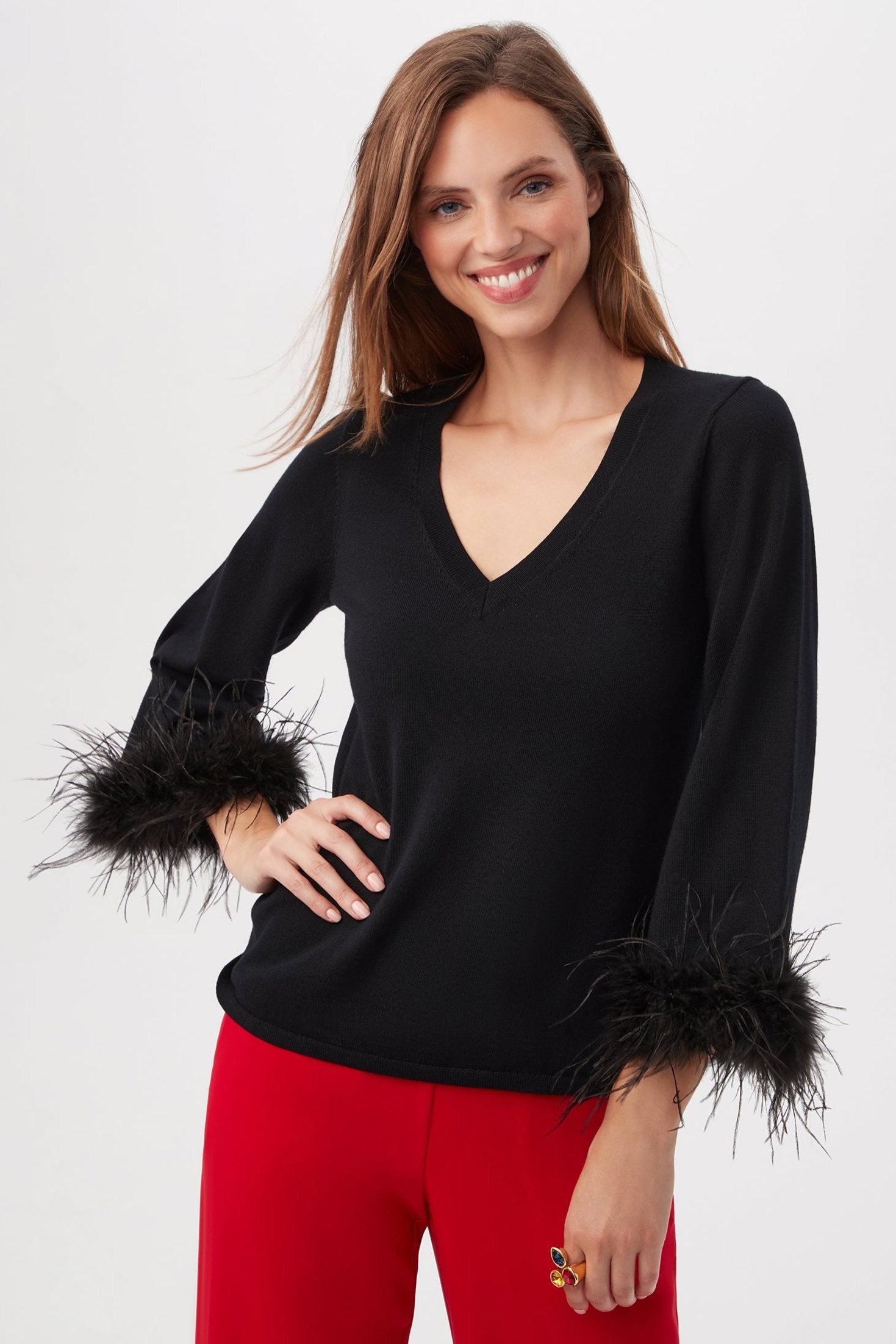 A smiling woman wears the Sassy Merino Wool Feather Trimmed Sweater in BLACK, featuring a V-neck and feathered cuffs, paired with bright red pants, standing with one hand on her hip against a plain light background.