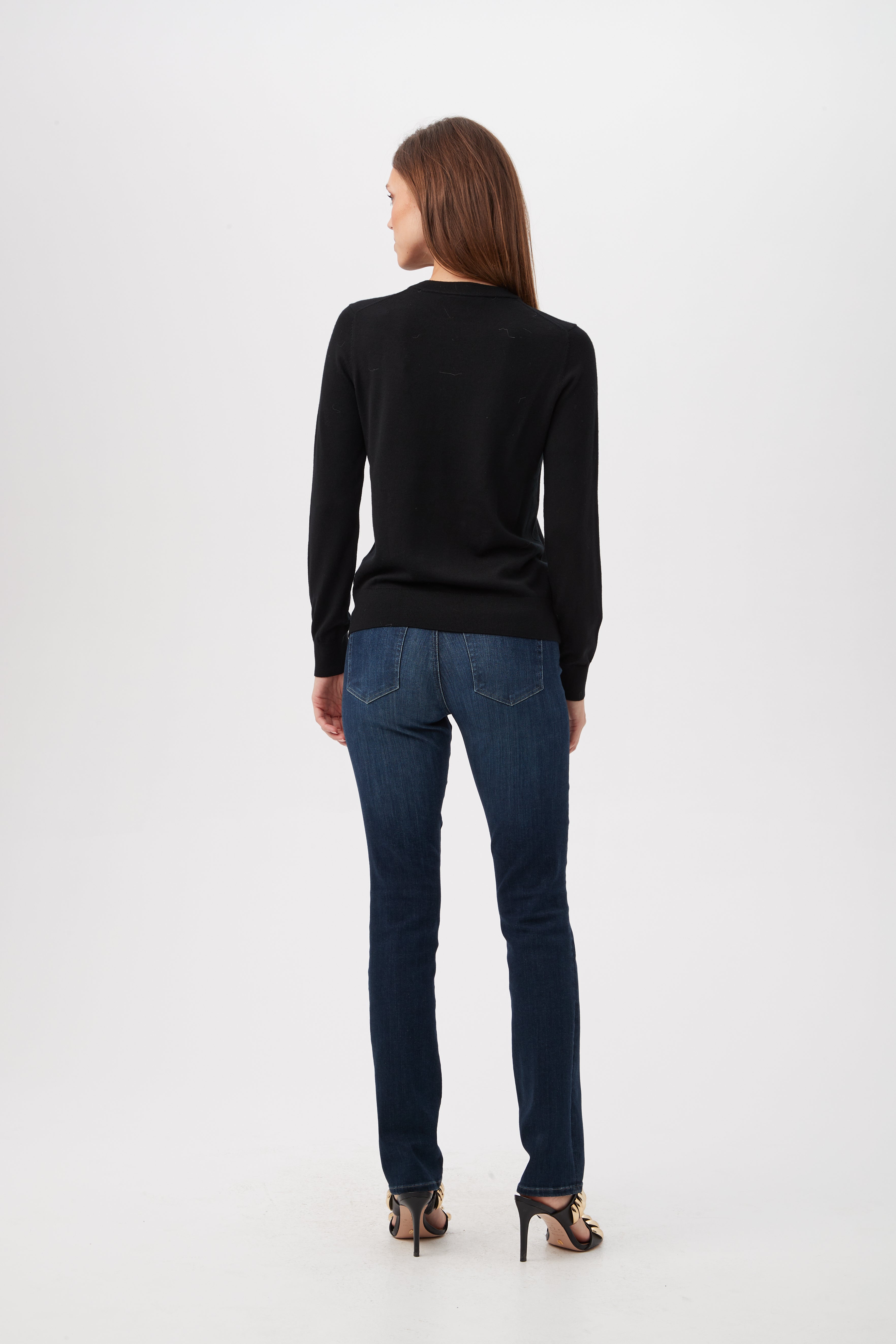 A woman with straight brown hair faces away, wearing the Queen B Merino Wool Sweater With Sequin Bee Detail, blue jeans, and black heeled sandals against a plain white background.