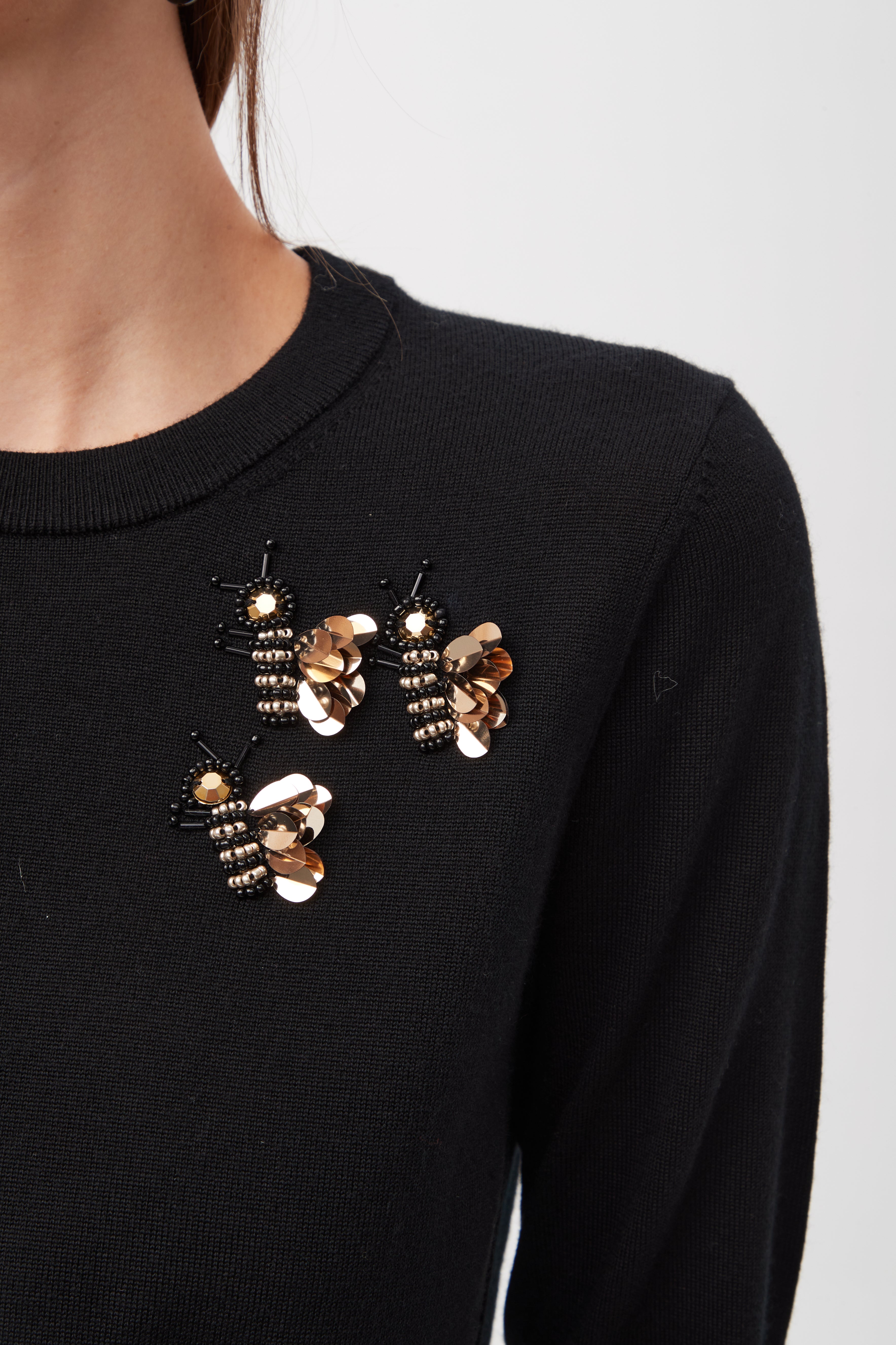A close-up shows a person wearing the Queen B Merino Wool Sweater With Sequin Bee Detail, featuring three gold and black beaded bee brooches pinned near the shoulder against a plain, light background.