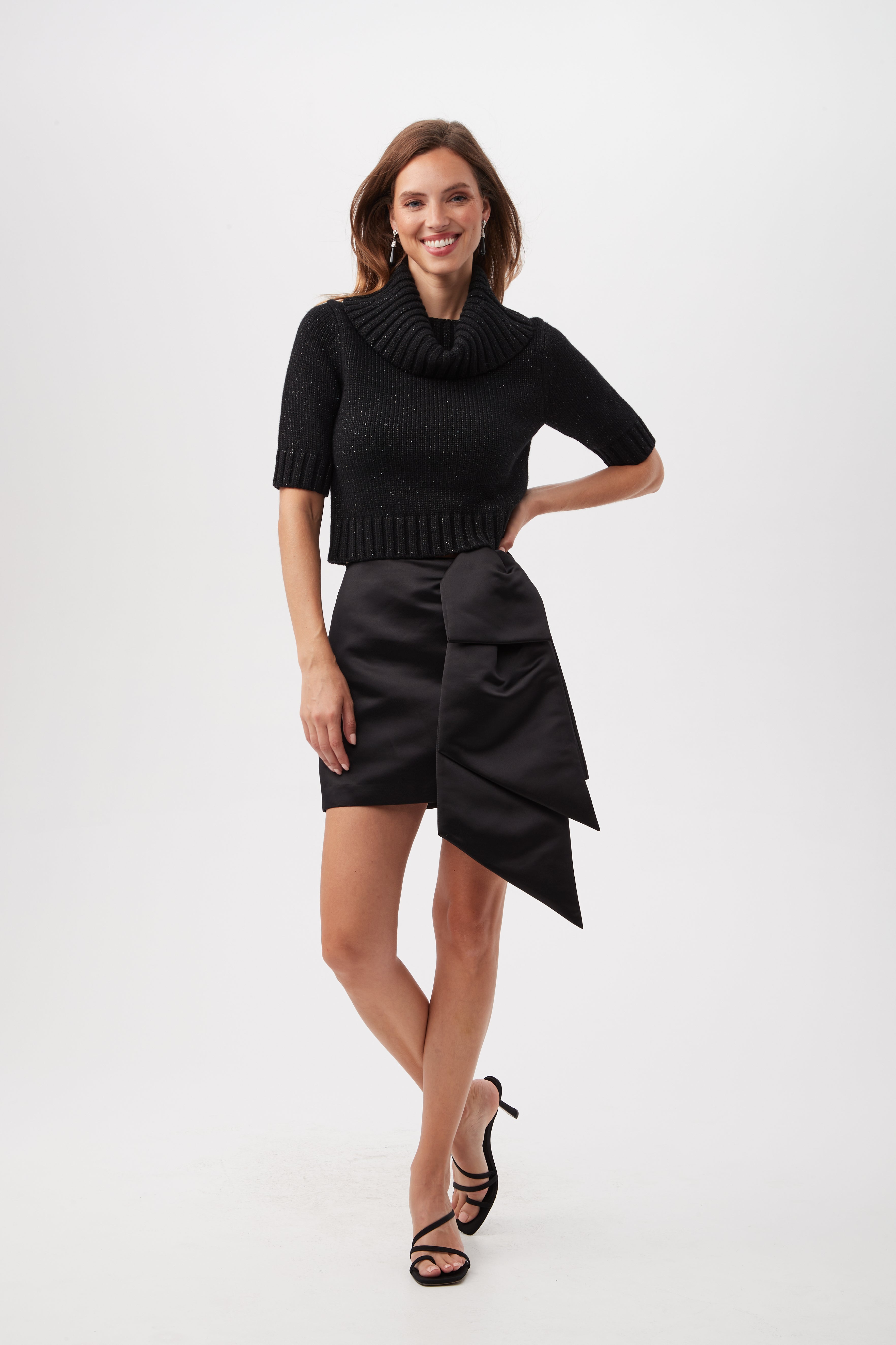 A woman smiles in the Titania Metallic Merino Wool Short Sleeve Sweater and a black satin mini skirt with a large bow, paired with black strappy high heels, standing against a plain white background.