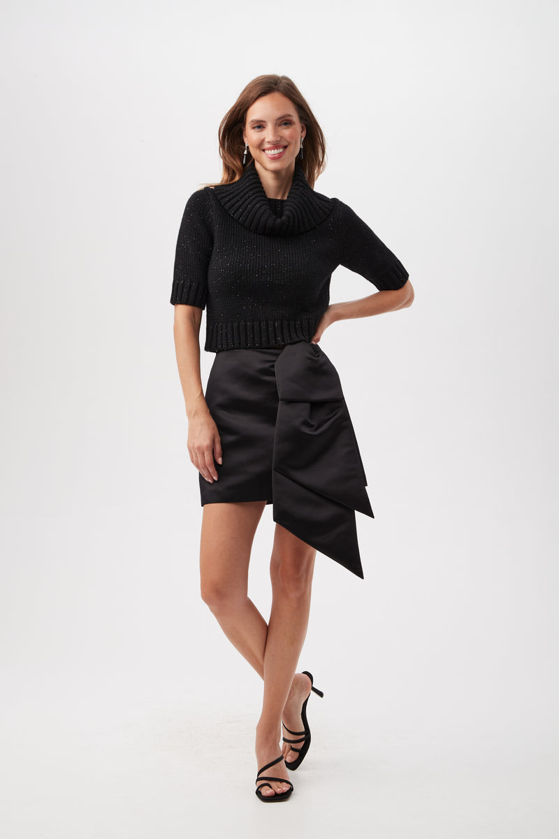 A woman smiles in the Titania Metallic Merino Wool Short Sleeve Sweater and a black satin mini skirt with a large bow, paired with black strappy high heels, standing against a plain white background.