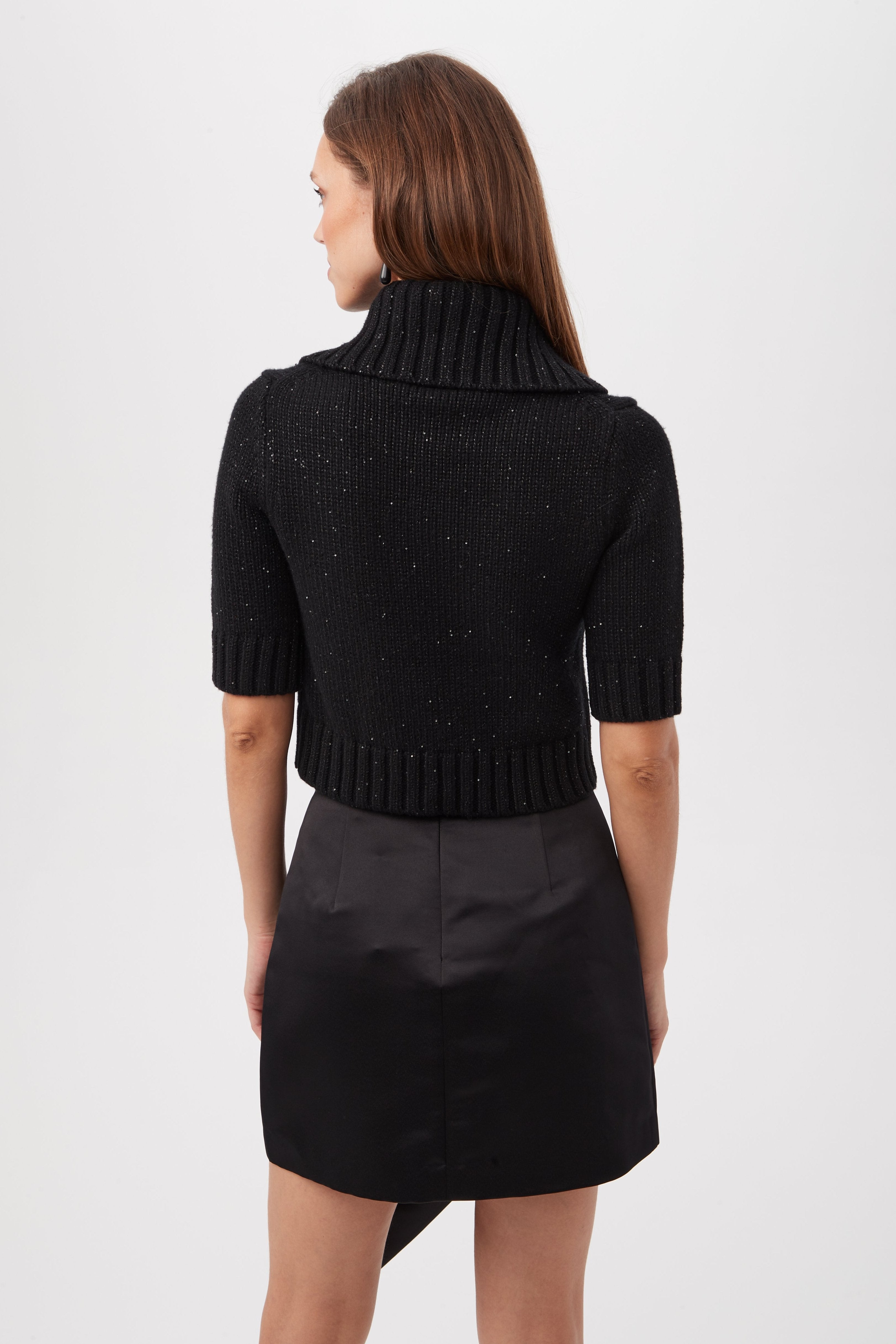 A woman with straight brown hair stands facing away, wearing the Titania Metallic Merino Wool Short Sleeve Sweater in black and a matching skirt against a plain light background.