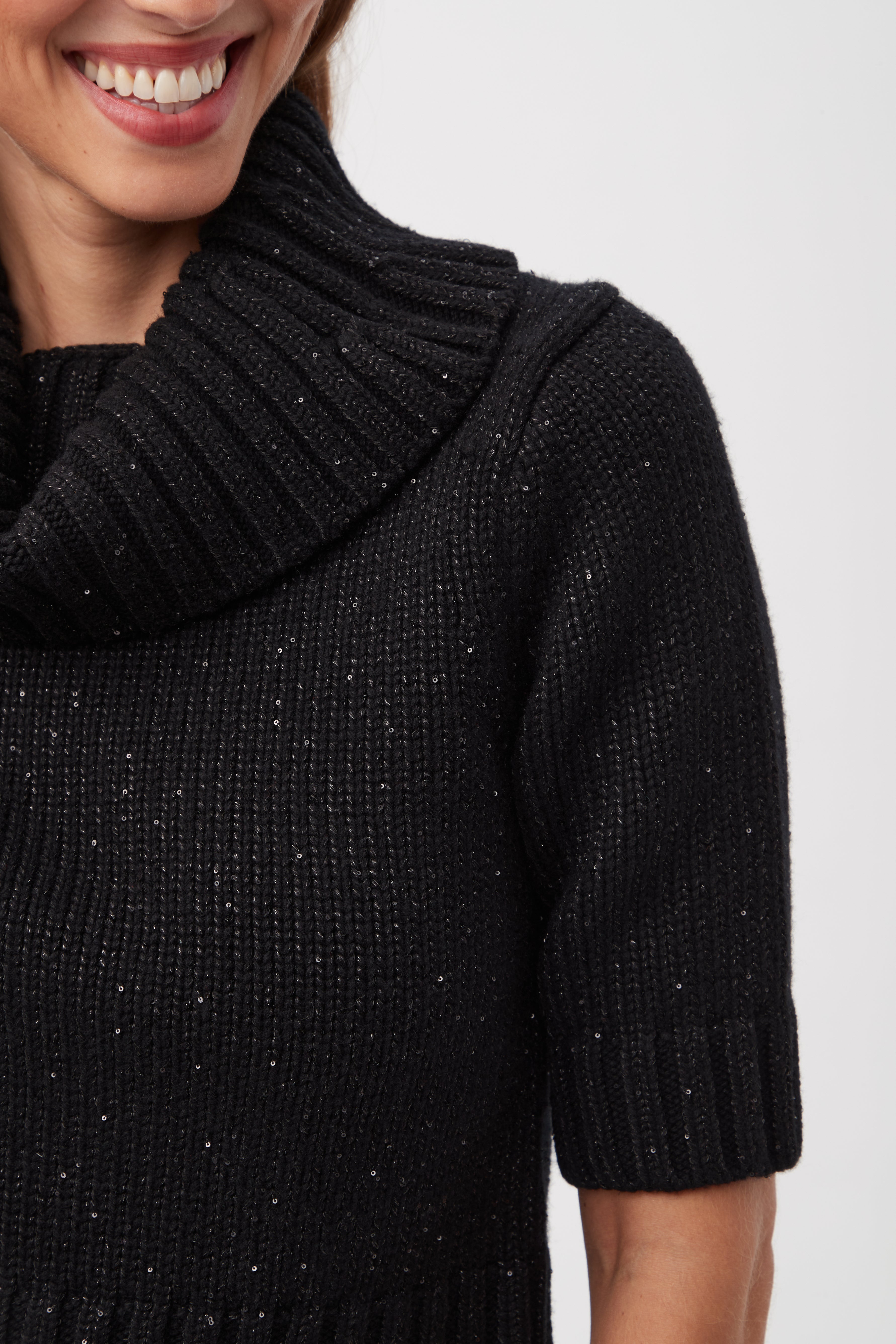 A woman smiles, showing her teeth, while wearing the Titania Metallic Merino Wool Short Sleeve Sweater—a ribbed black knit with a wide, folded turtleneck. Only her lower face, neck, and upper torso are visible.