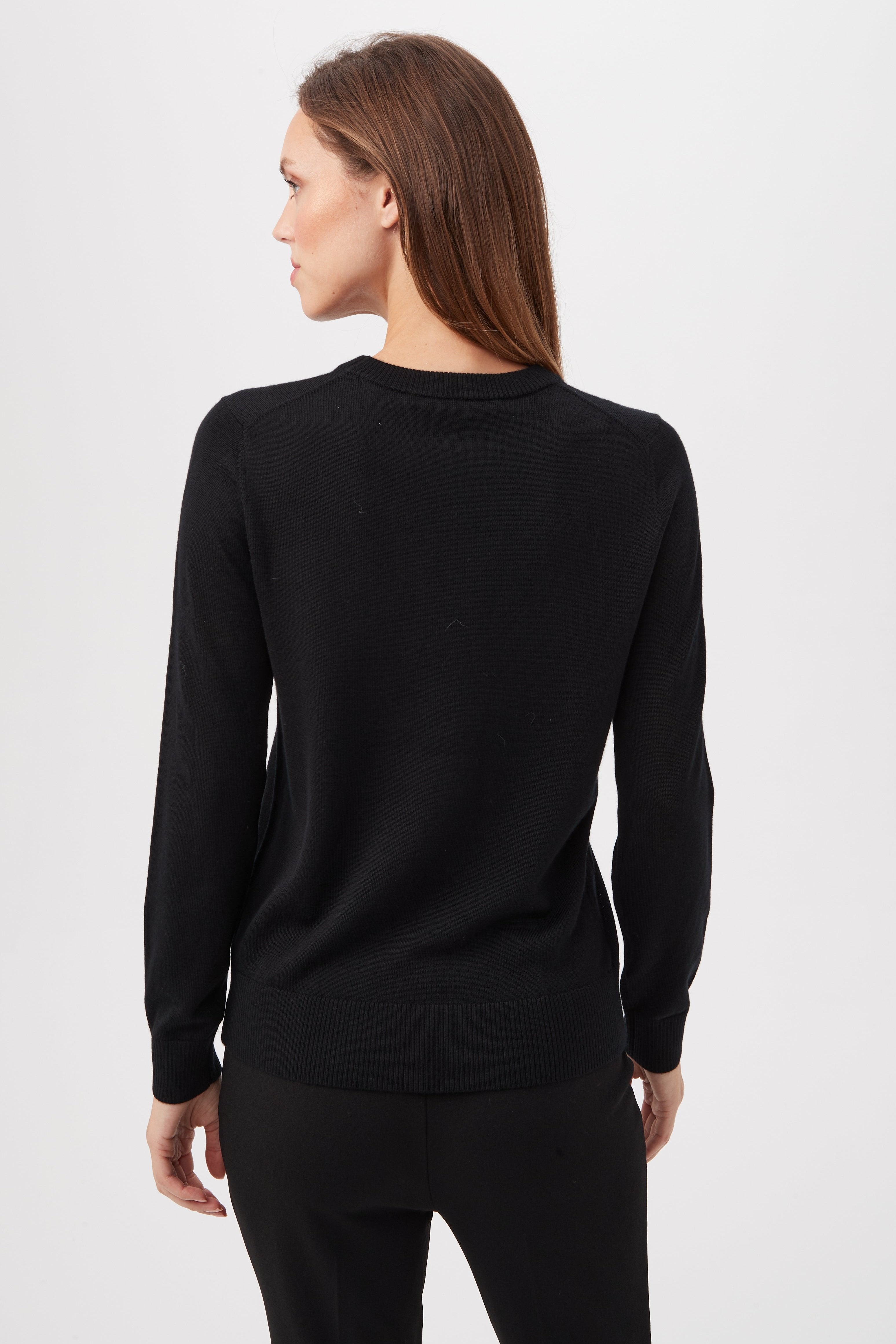 A woman with long brown hair faces away, wearing a black TT Martini Merino Wool Crewneck Sweater and black pants against a plain white background.