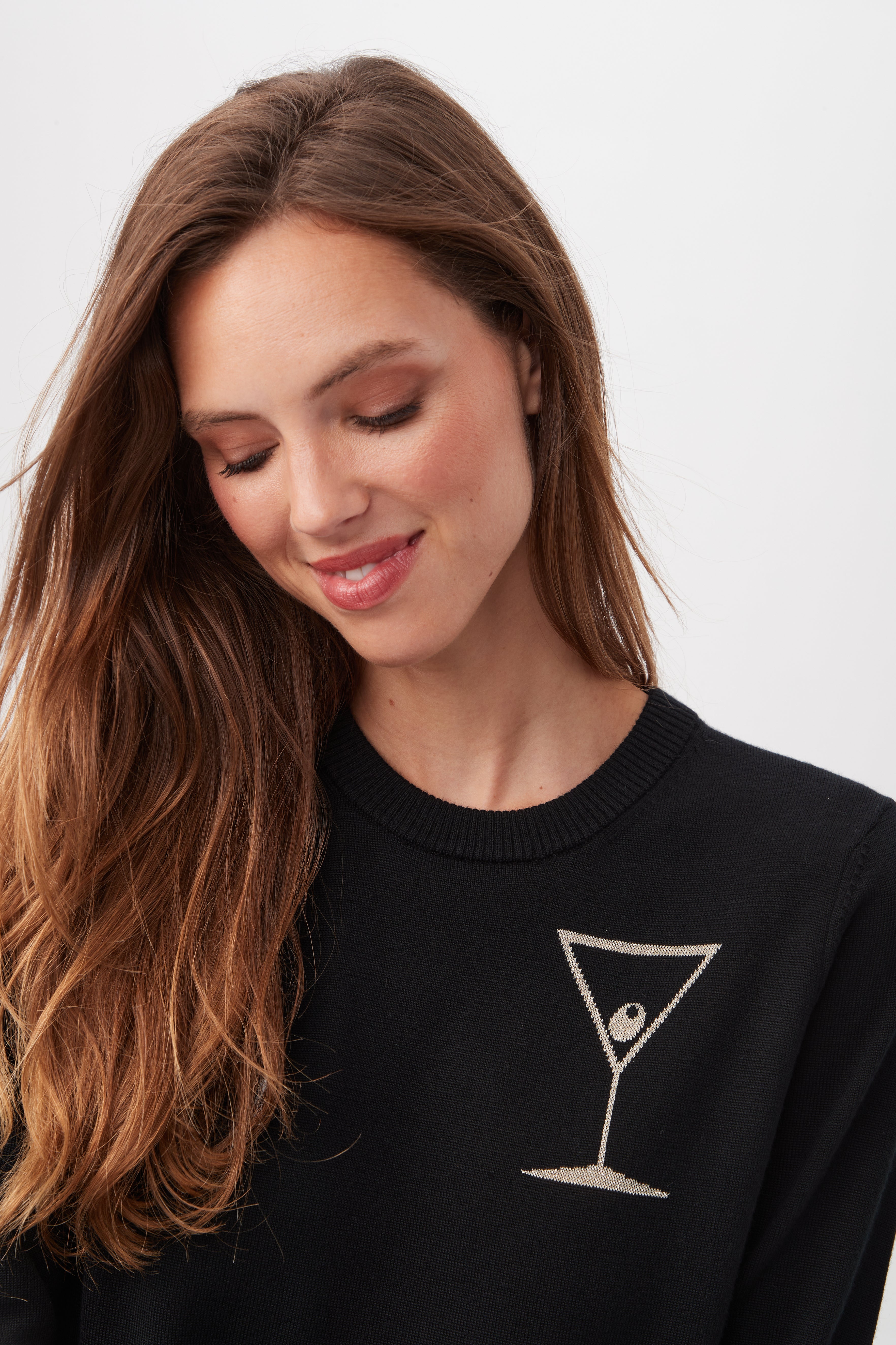 A woman with long brown hair smiles softly while looking down, wearing the TT Martini Merino Wool Crewneck Sweater featuring a small martini glass and olive graphic on the upper chest. The background is plain white.