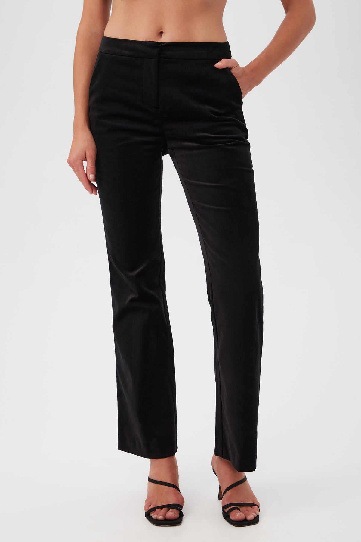 Someone stands against a white background wearing Westport Velveteen Bootcut Pants in black with one hand in their pocket, paired with black heeled sandals and a visible midriff.