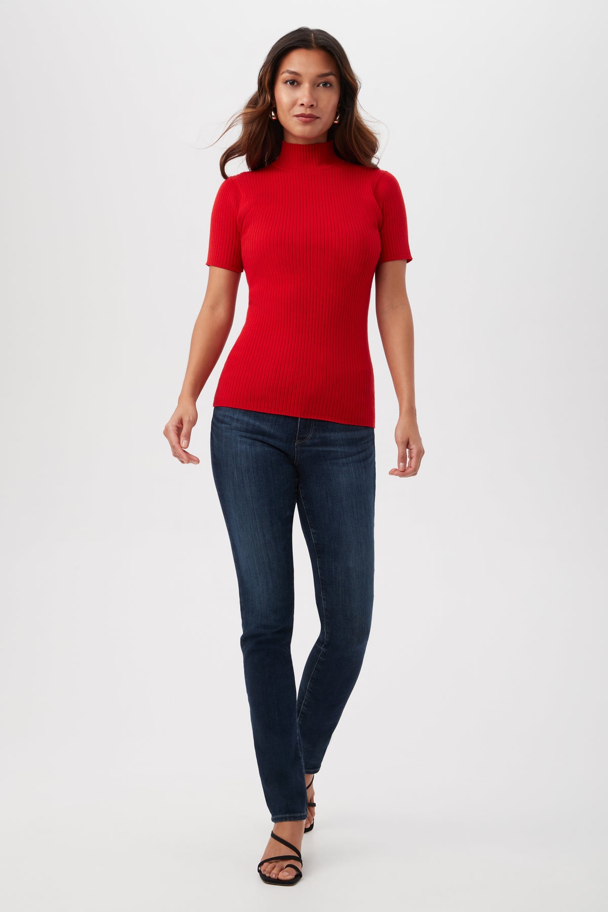A woman with long hair wears the Armeni Merino Wool Short Sleeve Mockneck Top in red, paired with dark blue skinny jeans and black strappy sandals. She stands against a plain white background, looking forward with a neutral expression.