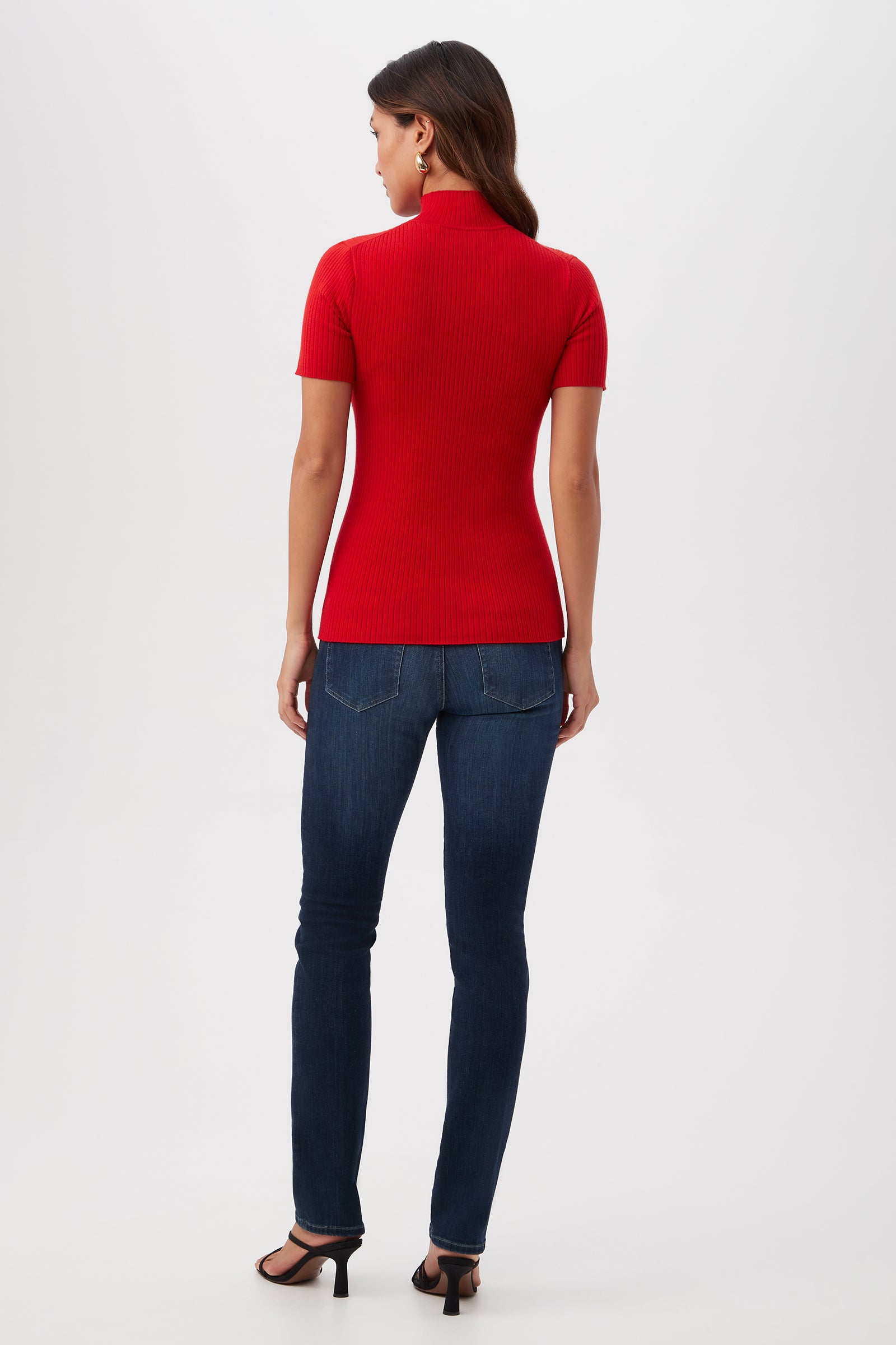 A woman faces away, wearing the Armeni Merino Wool Short Sleeve Mockneck Top in red, paired with dark blue skinny jeans and black heeled sandals, set against a plain white background.