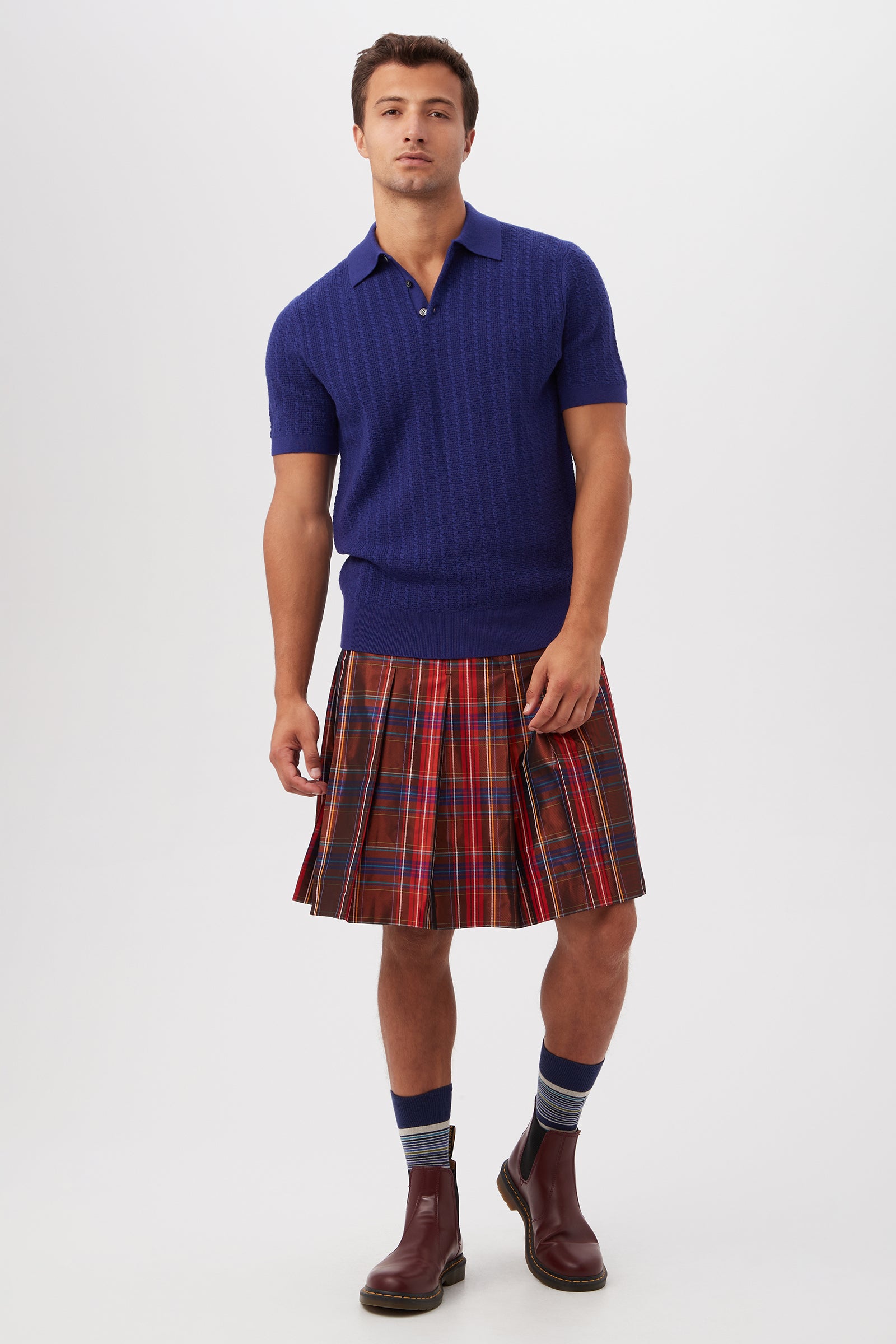 A man with short hair and a relaxed posture stands against a plain background wearing the Asher Short-Sleeve Polo - Merino Wool in blue, a red tartan kilt, blue striped socks, and brown boots.