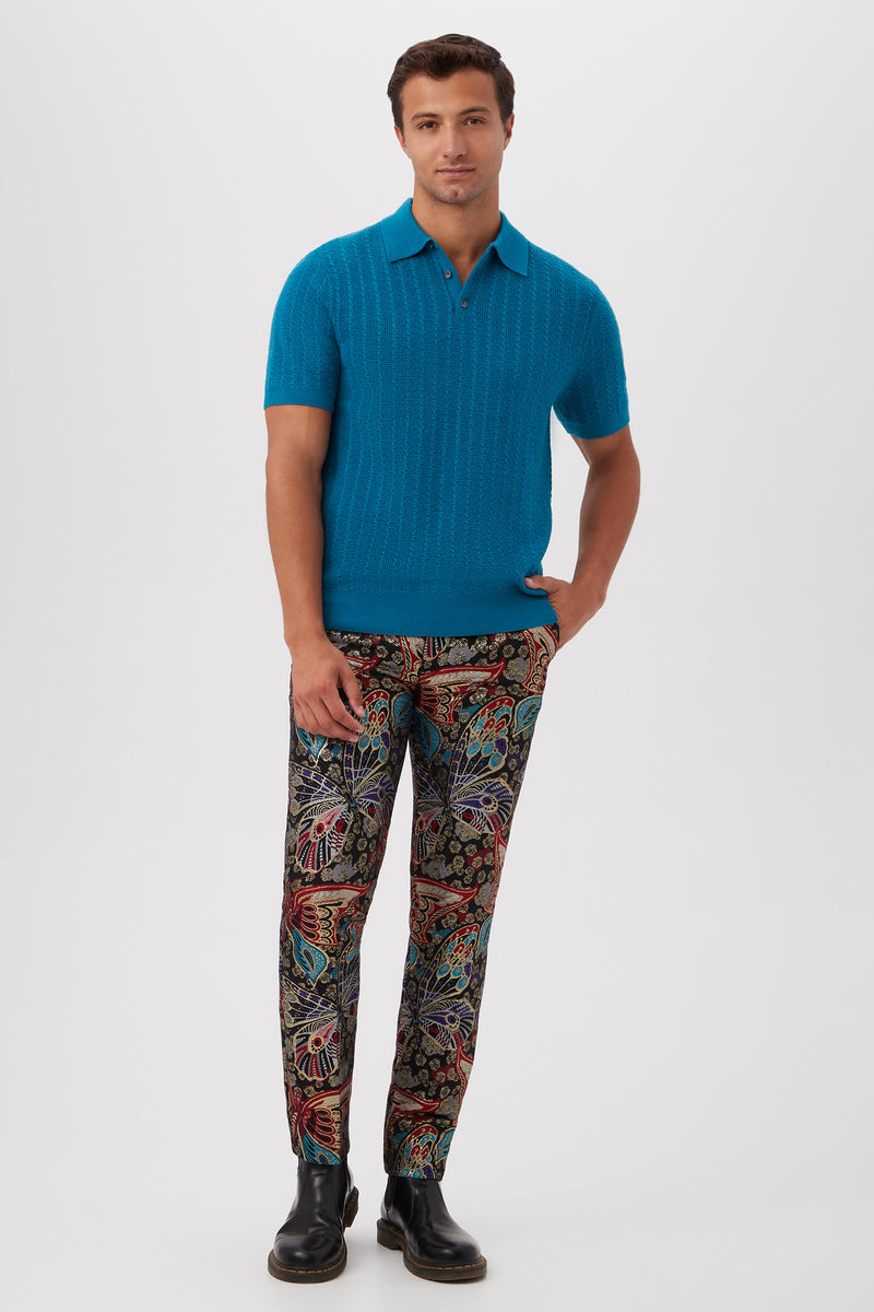 A man poses against a plain background wearing the Asher Short-Sleeve Polo in bright blue merino wool, colorful floral and abstract patterned trousers, and black boots.