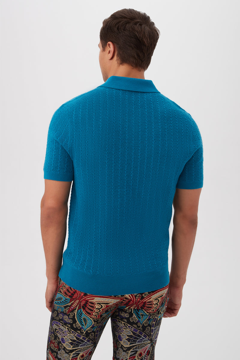 A man faces away, wearing the Asher Short-Sleeve Polo in textured blue merino wool and colorful paisley-patterned pants, set against a plain white background.