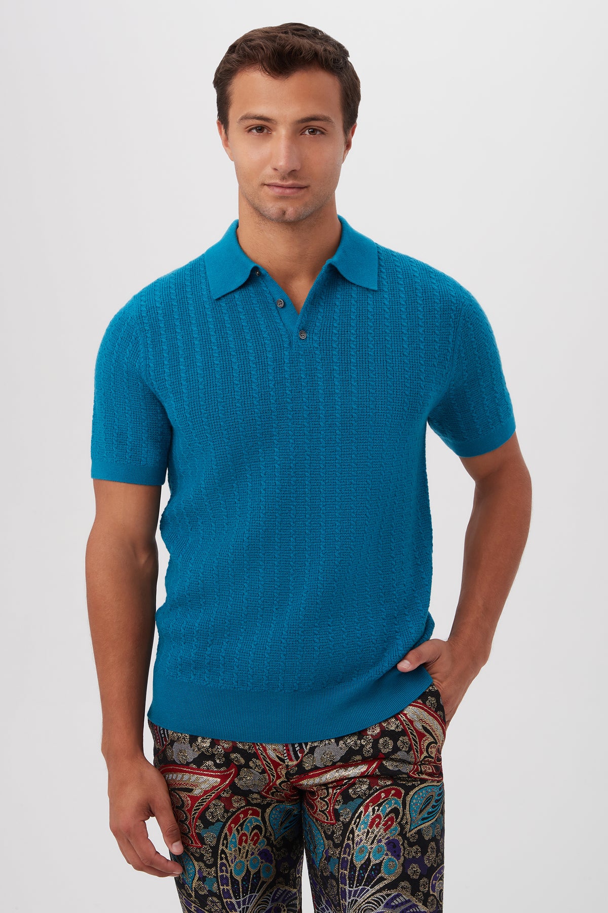 A man with short brown hair wears the Asher Short-Sleeve Polo in Taboo Teal merino wool and colorful patterned pants. He stands against a plain light background, with one hand in his pocket and a neutral expression.