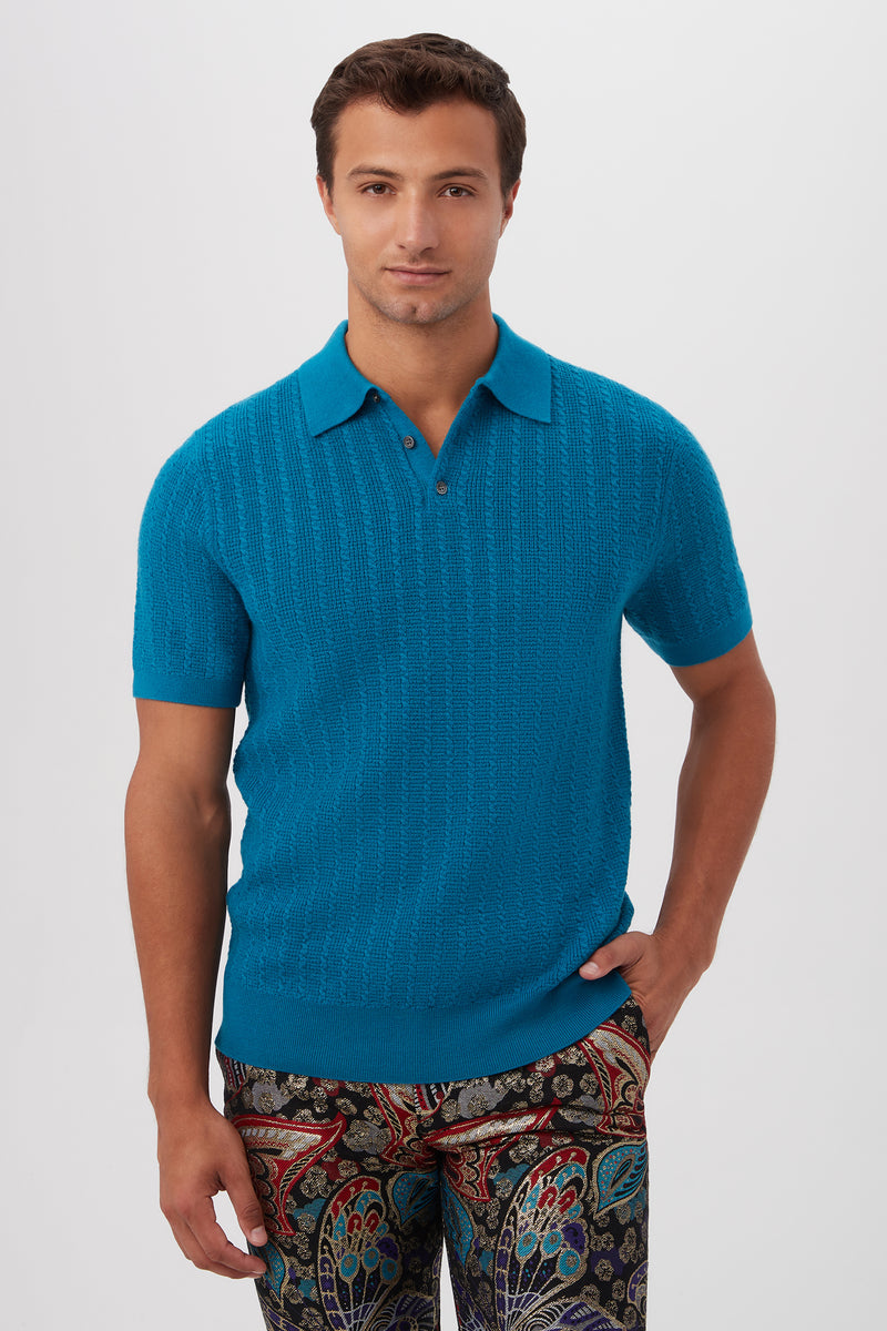 A man with short brown hair wears the Asher Short-Sleeve Polo in Taboo Teal merino wool and colorful patterned pants. He stands against a plain light background, with one hand in his pocket and a neutral expression.