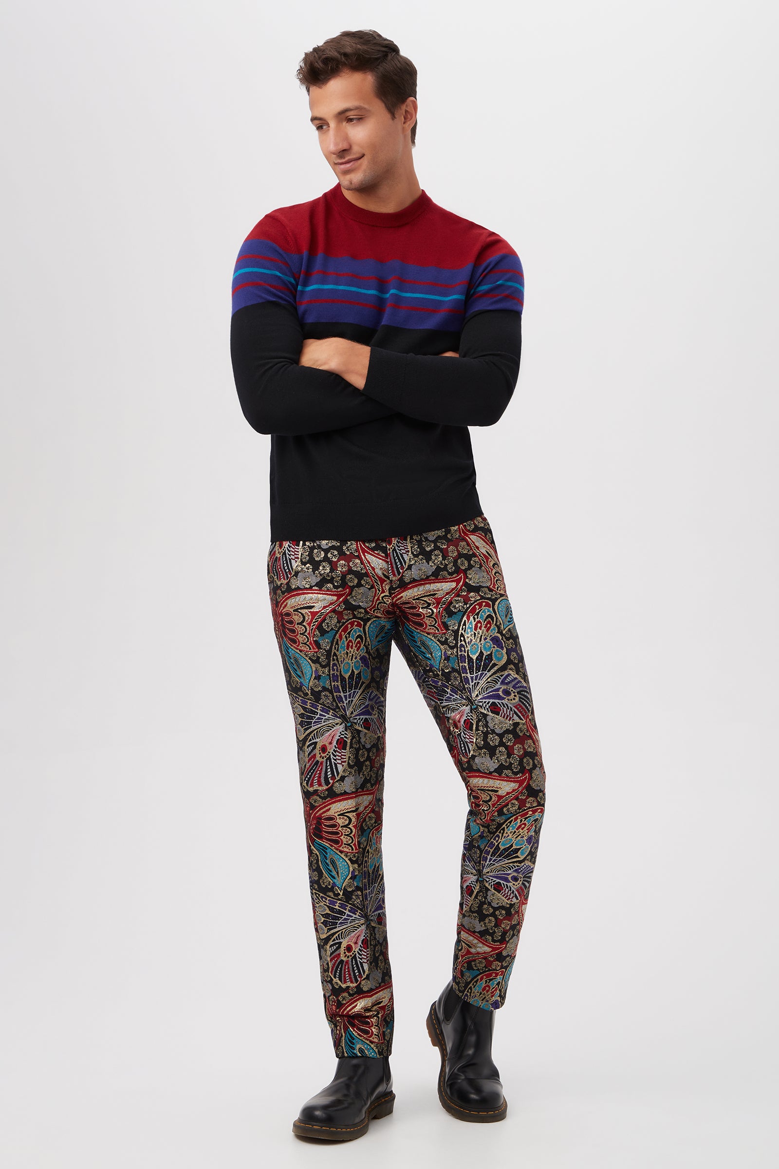 A man stands with arms crossed, wearing the Romero Crewneck Sweater in black and red with blue stripes, paired with colorful floral and paisley patterned pants and black boots. He looks to the side, smiling.