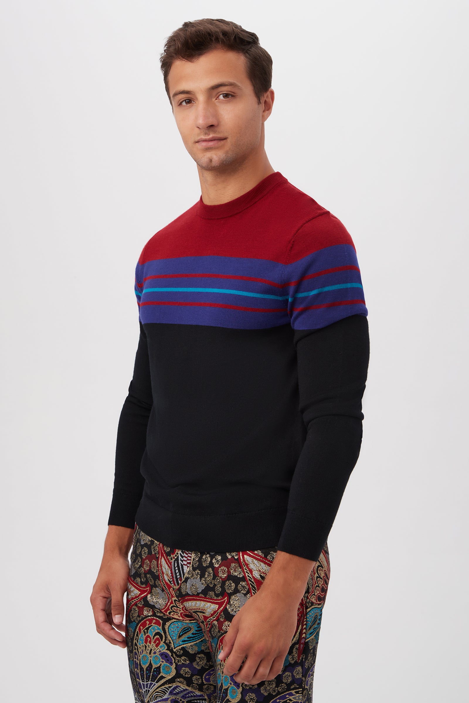 A man poses against a white background wearing the Romero Crewneck Sweater, a black knit with red and blue horizontal stripes on the chest and shoulders, paired with patterned pants featuring colorful, intricate designs.