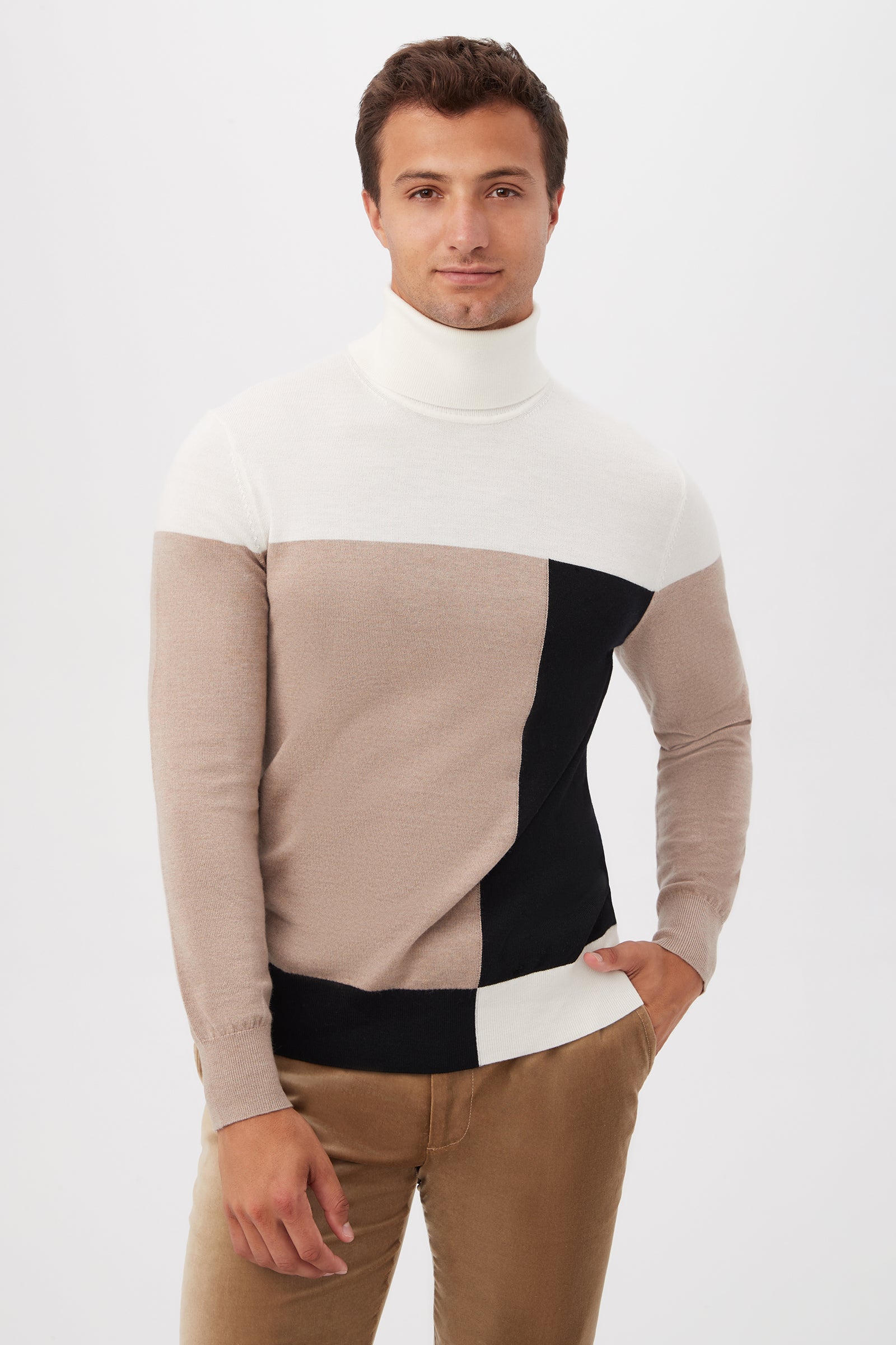 A man poses against a plain white background wearing the Callisto Turtleneck Sweater in Multi, featuring white, beige, and black color-block sections, with one hand in his pocket.