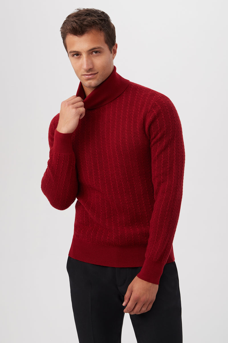 A man stands against a plain, light background, wearing the Woodrow Turtleneck Sweater in textured red Merino wool and black pants. He holds the sweaters collar with one hand, looking slightly to the side.