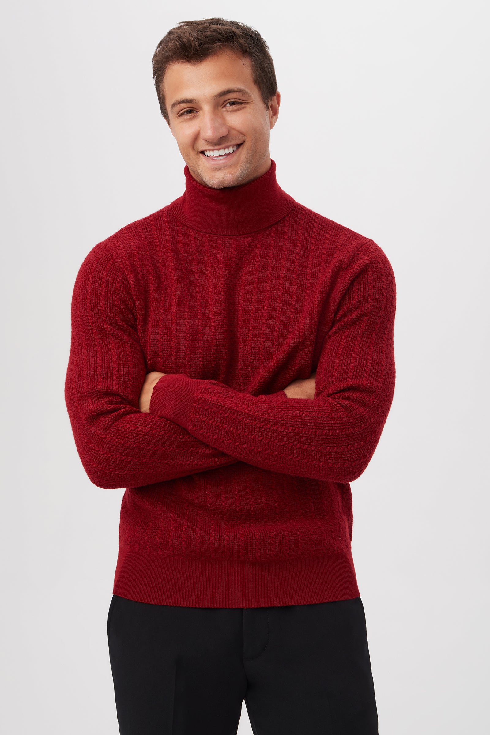 A young man with short brown hair smiles with arms crossed, wearing the Woodrow Turtleneck Sweater in merino wool and black pants, standing against a plain light gray background.