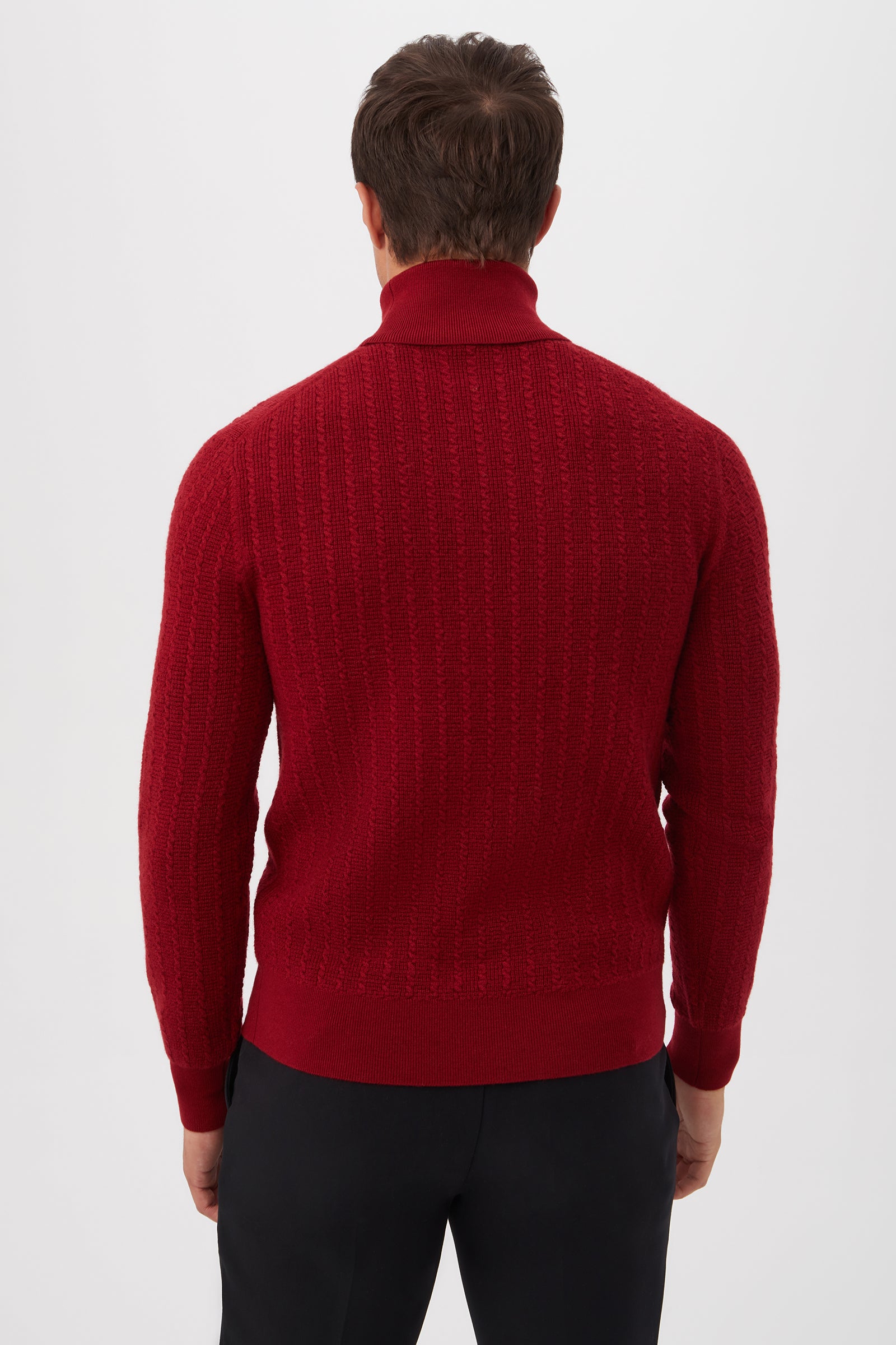 A man stands with his back to the camera, wearing the red Woodrow Turtleneck Sweater made from merino wool and black pants, set against a plain white background.