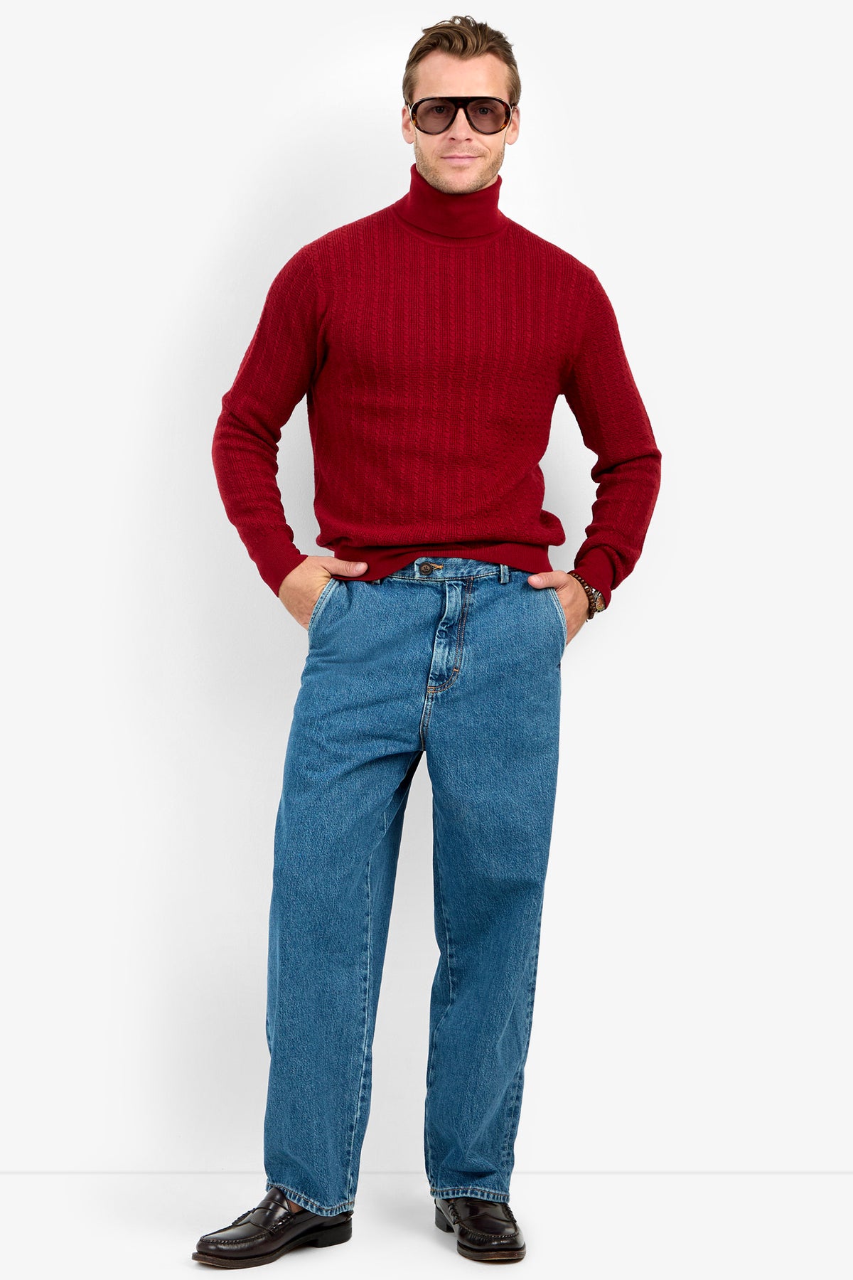 Man wears the Woodrow Turtleneck Sweater - Merino Wool in red, with blue jeans, black shoes, and sunglasses, standing hands-in-pockets against a plain white background.