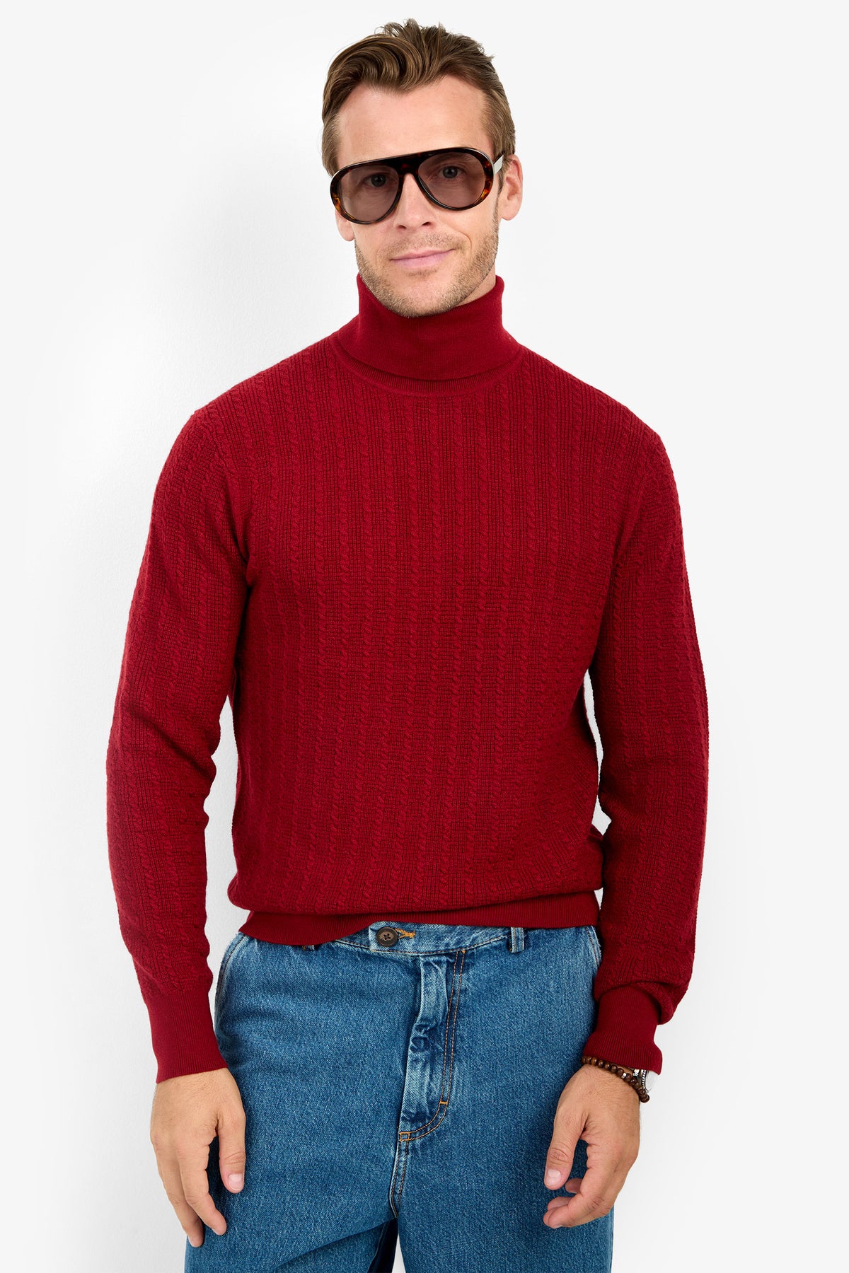 A person wearing the Woodrow Turtleneck Sweater - Merino Wool in red, blue jeans, and dark sunglasses stands facing forward against a plain white background with a neutral expression.