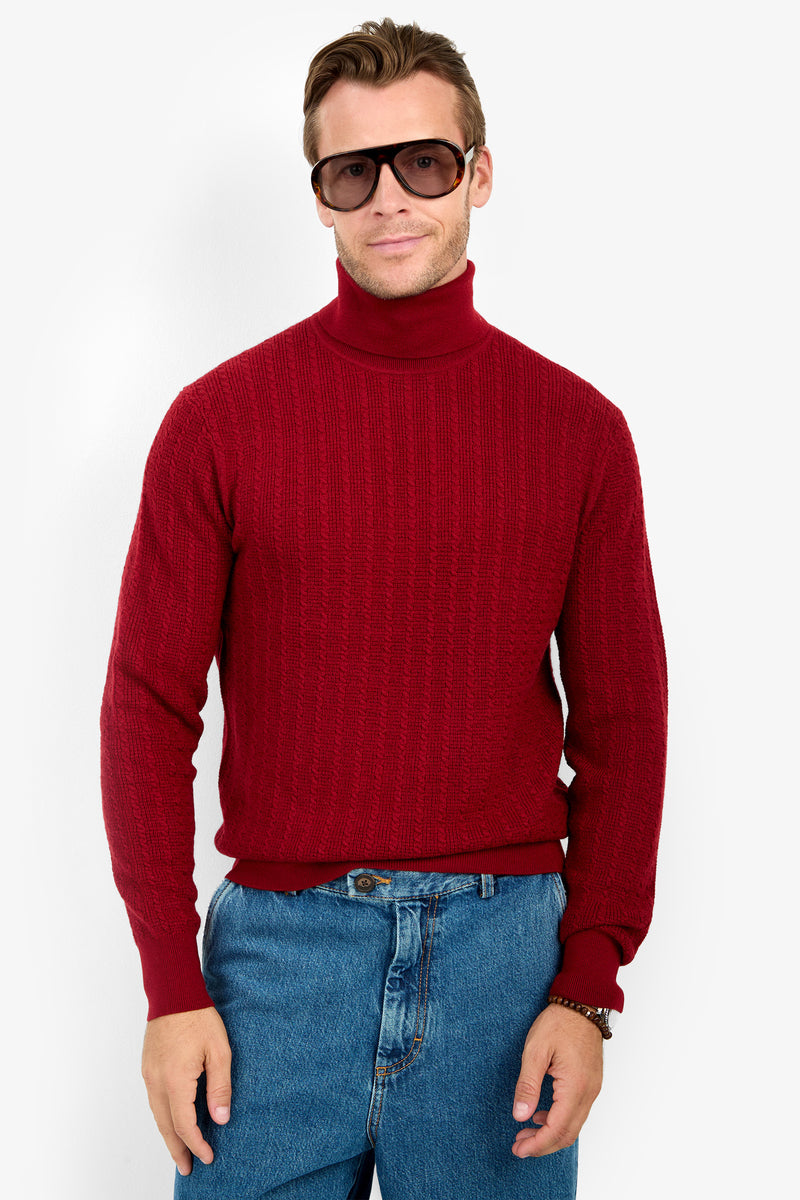 A person wearing the Woodrow Turtleneck Sweater - Merino Wool in red, blue jeans, and dark sunglasses stands facing forward against a plain white background with a neutral expression.