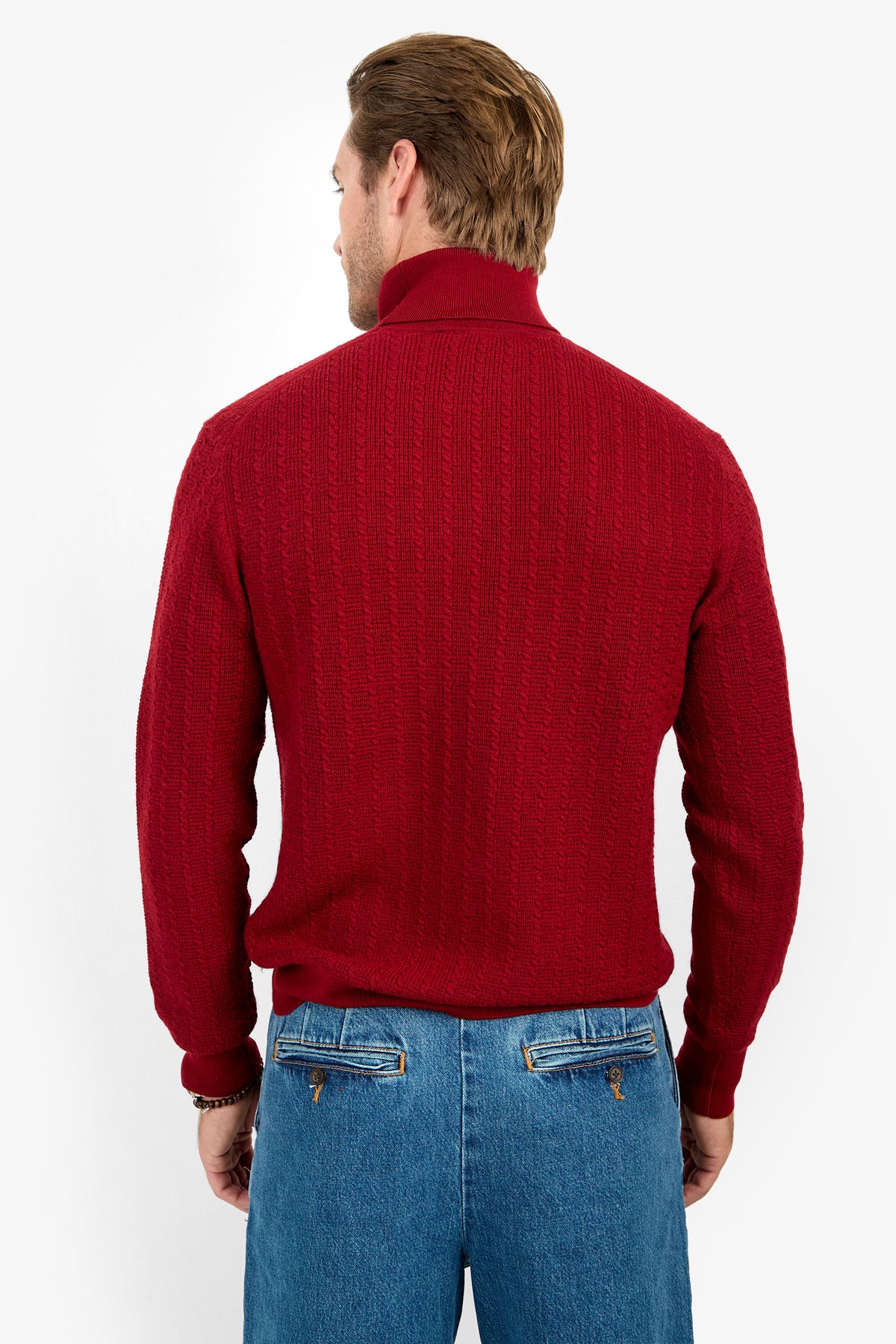 A man faces away, wearing the Woodrow Turtleneck Sweater - Merino Wool in red and blue jeans with brown leather back pocket accents, set against a plain white background.