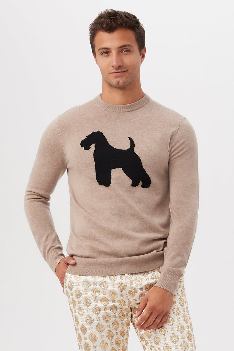 A young man wears the taupe Weston Airedale Terrier Sweater in merino wool, featuring a bold black dog silhouette on the front, paired with white pants with a gold geometric pattern, standing against a plain white background.