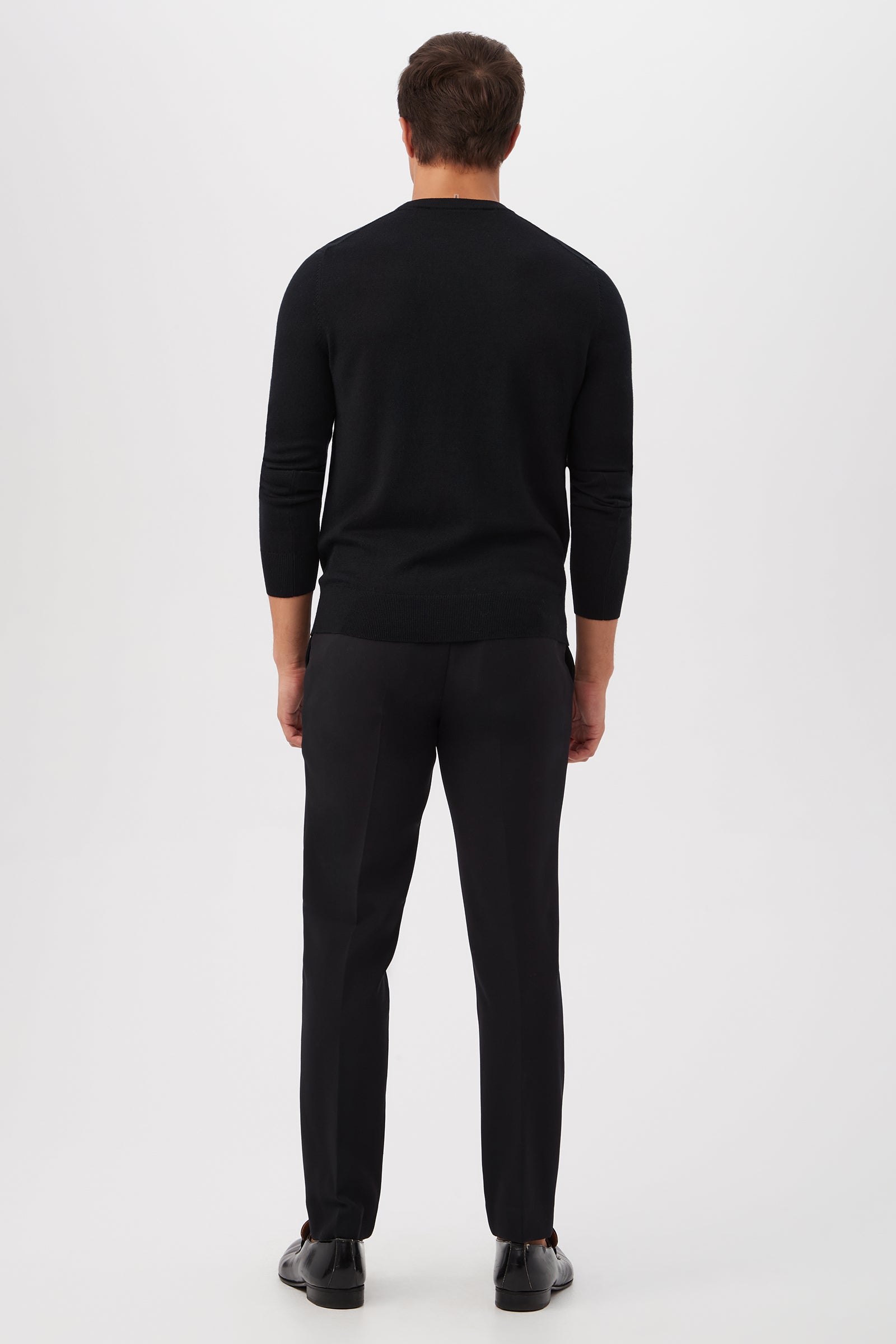 A man stands facing away from the camera, wearing the Mt Martini Long-Sleeve Sweater - Merino Wool in black, along with matching black trousers and shoes, set against a plain white background.