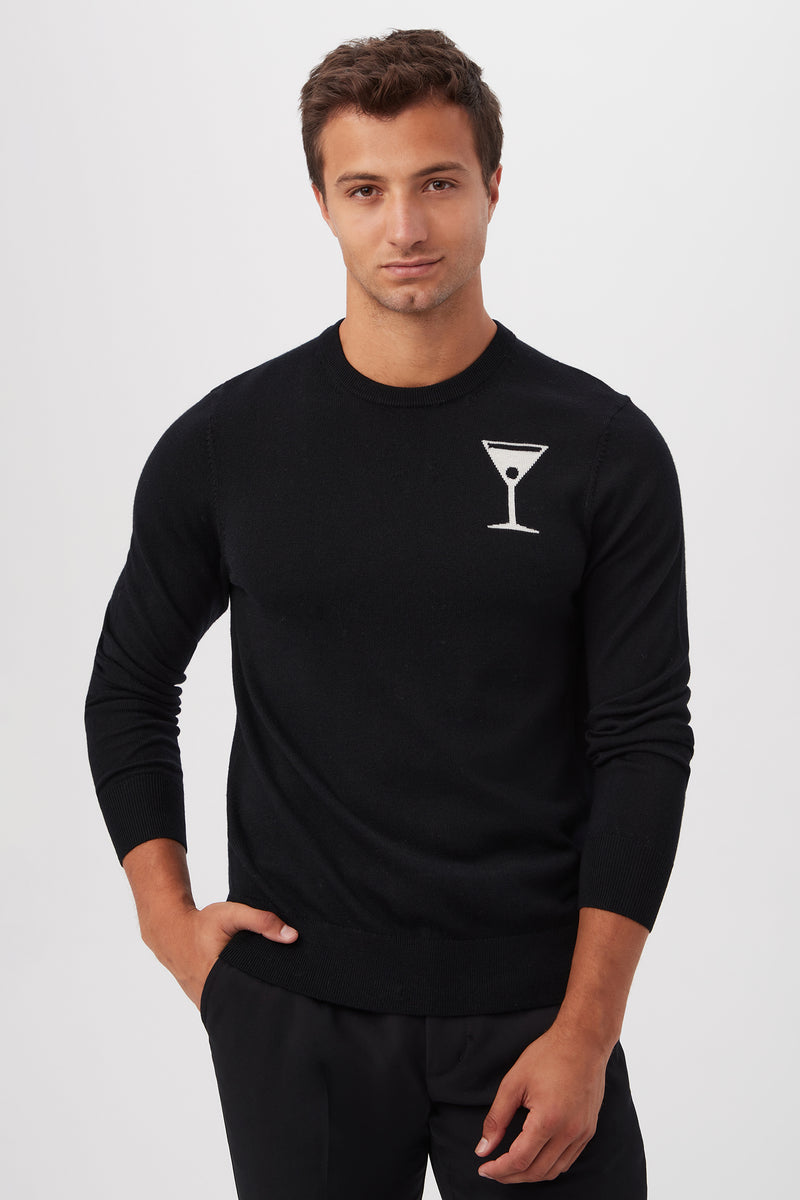 A man wears the Mt Martini Long-Sleeve Sweater - Merino Wool, featuring a white martini glass graphic on the left chest, as he stands against a plain white background with a neutral expression and one hand in his pocket.