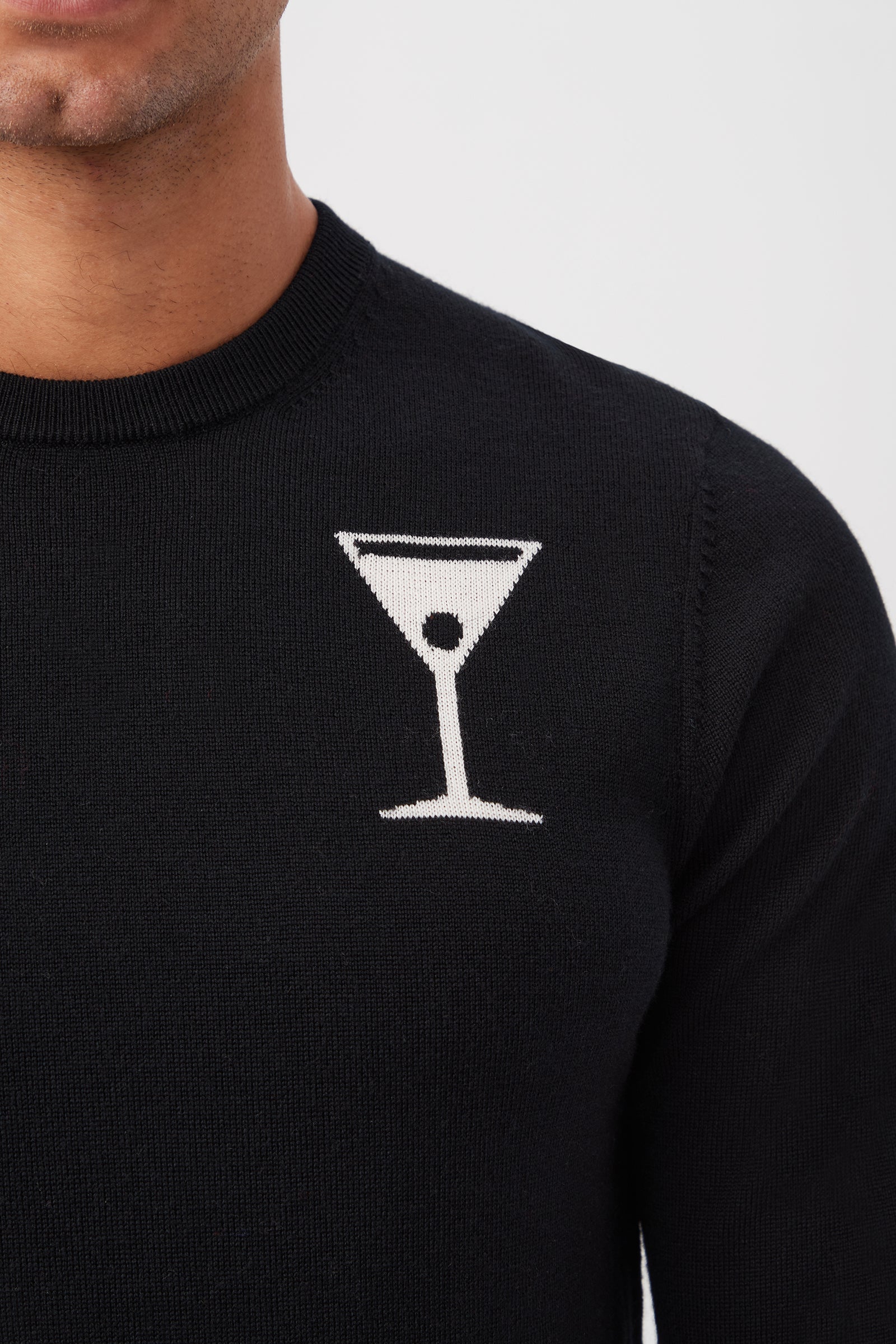 A person is seen from the lower face down, wearing the Mt Martini Long-Sleeve Sweater—made of merino wool—in black with a white martini glass graphic on the left chest.