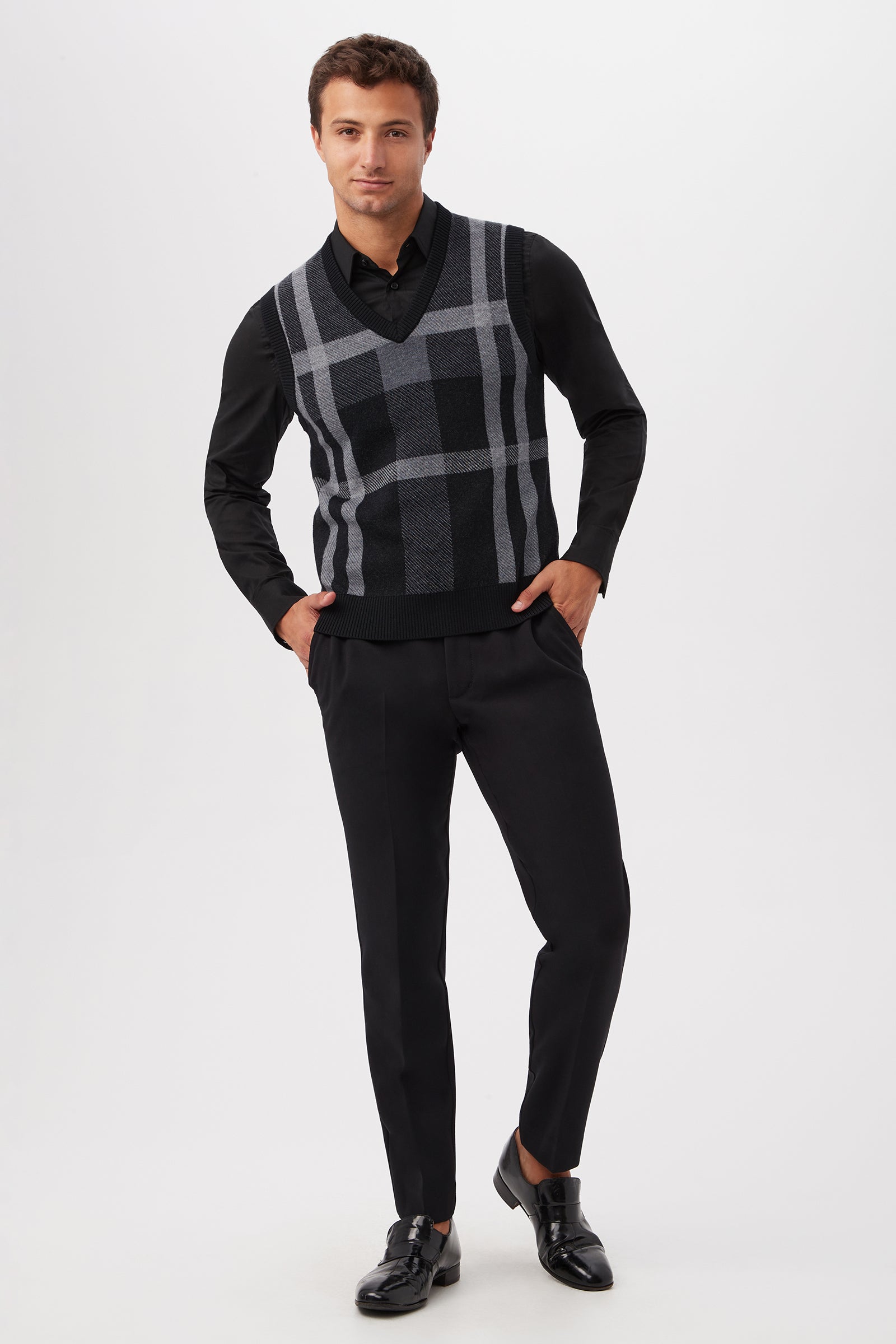 A man stands against a white background, wearing a black button-up shirt, black pants, black dress shoes, and the Neso Plaid Knit Sweater Vest - Merino Wool in black and gray plaid. He has one hand in his pocket and smiles slightly.